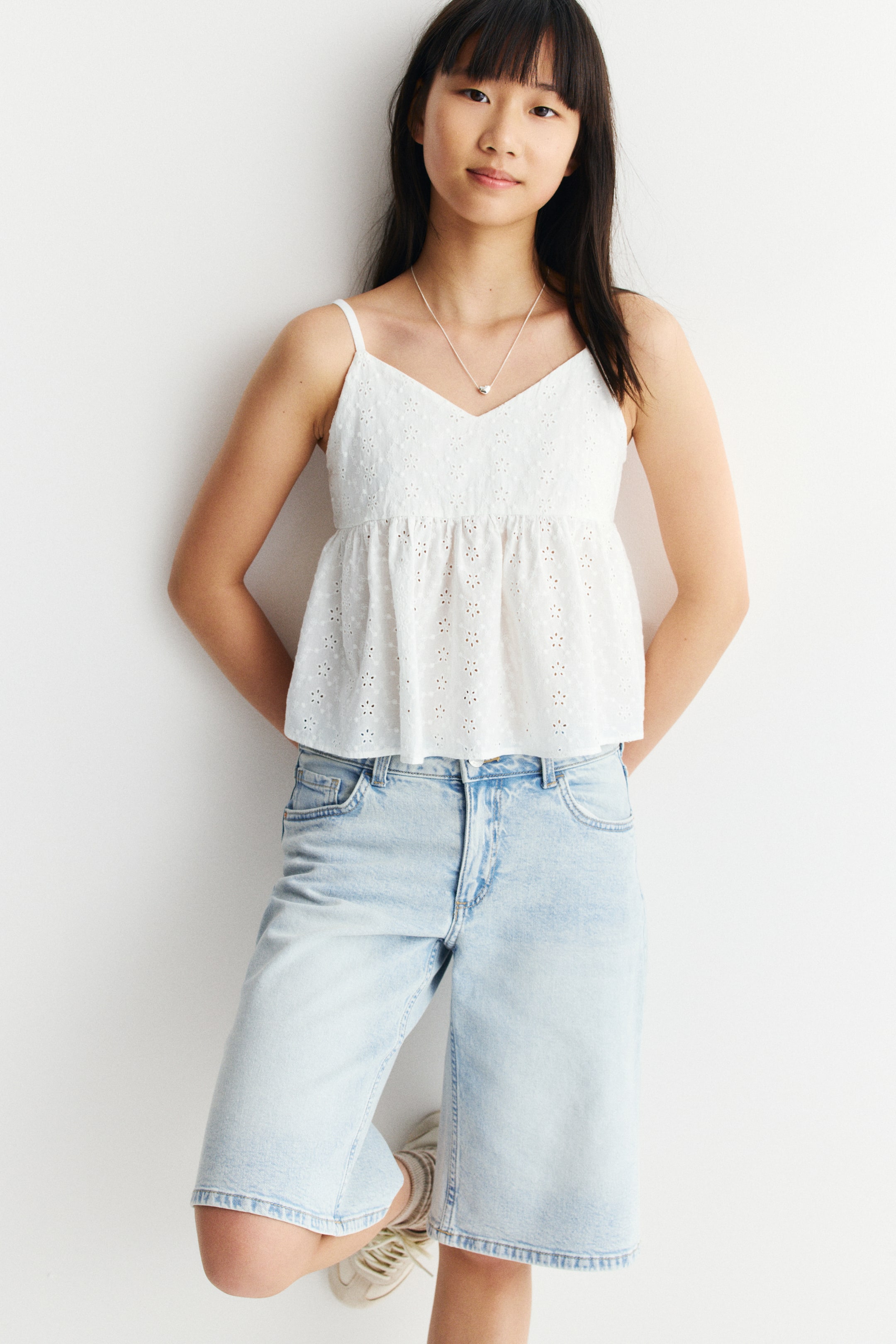 View larger image: A white eyelet camisole top is showcasing a V-neckline and thin spaghetti straps, complemented by a gathered peplum hem. It is paired with light wash denim Bermuda shorts, which exhibit a relaxed wide-leg fit and prominent front pockets.