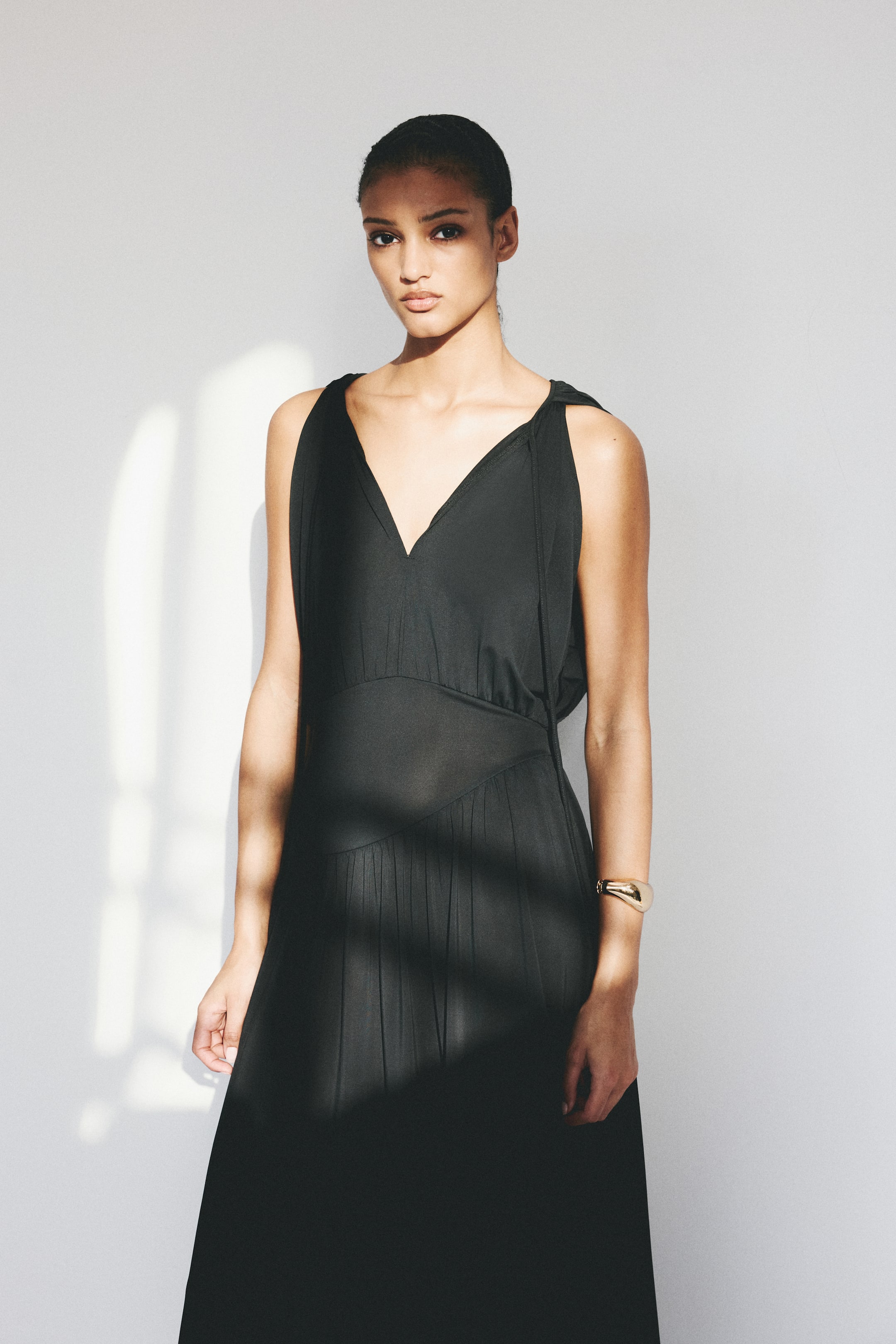 Grösseres Bild ansehen: Ein schwarzes ärmelloses Kleid wird präsentiert, das mit einem tiefen V-Ausschnitt und breiten, drapierten Schulterträgern ausgestattet ist. Das Kleid zeigt ein eng anliegendes Oberteil und ein definiertes Taillenband, das in einen fließenden Rock mit sanften Falten übergeht.