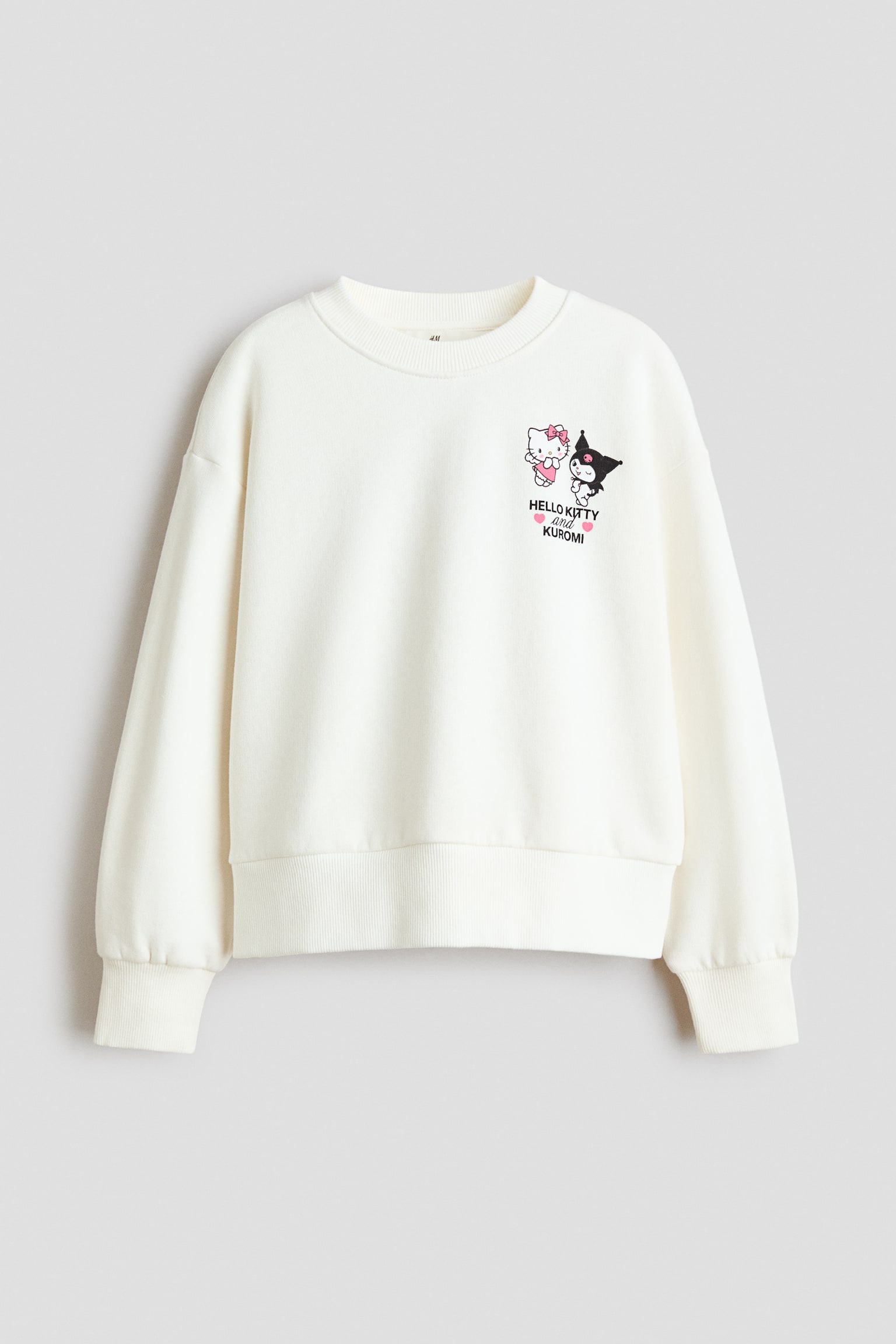 Una sudadera de corte holgado y color crema se muestra de frente, con escote redondo acanalado, hombros caídos y mangas largas con puños y bajo acanalados. En el pecho izquierdo presenta un pequeño estampado gráfico de los personajes Hello Kitty y Kuromi con el texto "HELLO KITTY and KUROMI" debajo.