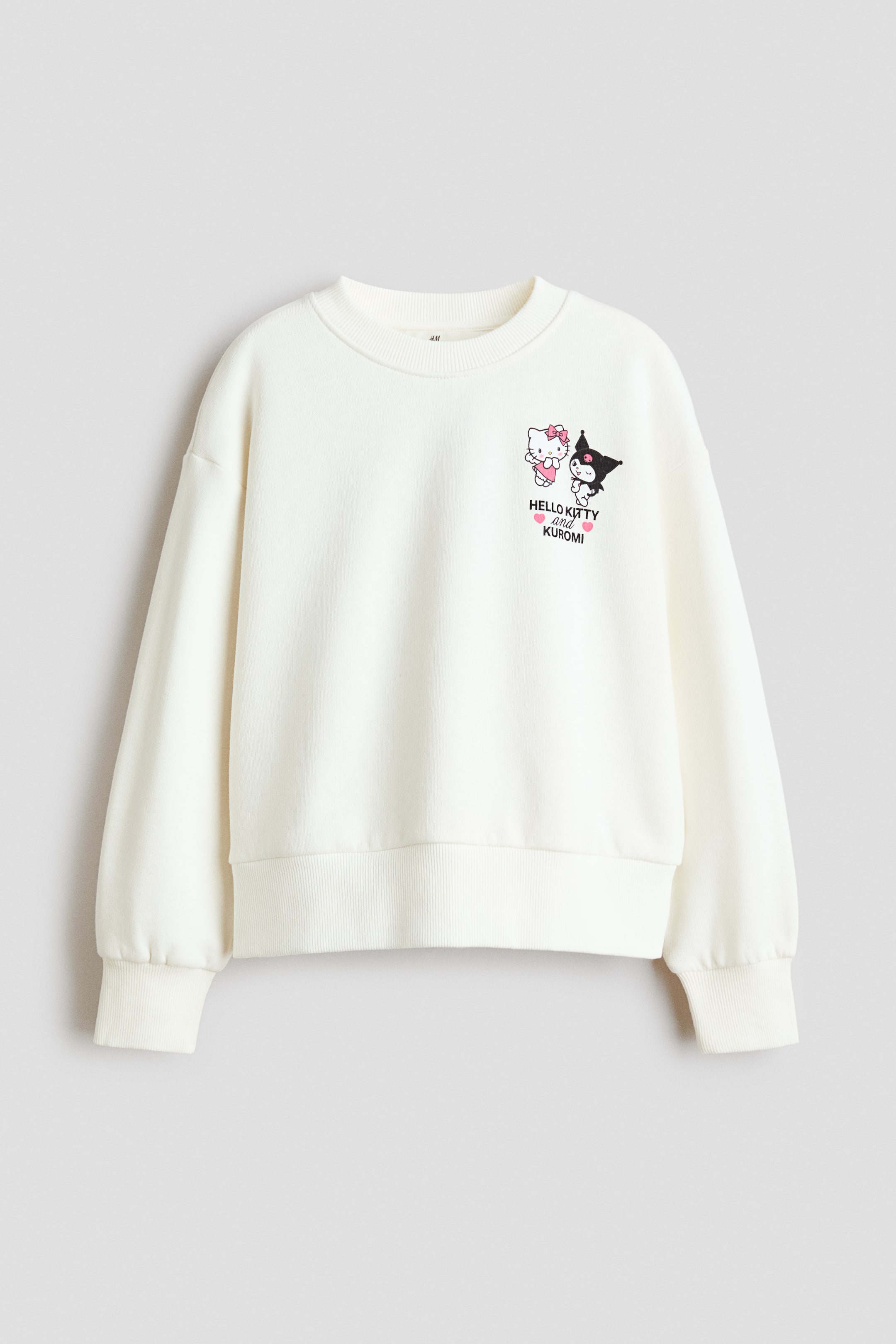 Ver imagen más grande: Una sudadera de corte holgado y color crema se muestra de frente, con escote redondo acanalado, hombros caídos y mangas largas con puños y bajo acanalados. En el pecho izquierdo presenta un pequeño estampado gráfico de los personajes Hello Kitty y Kuromi con el texto "HELLO KITTY and KUROMI" debajo.