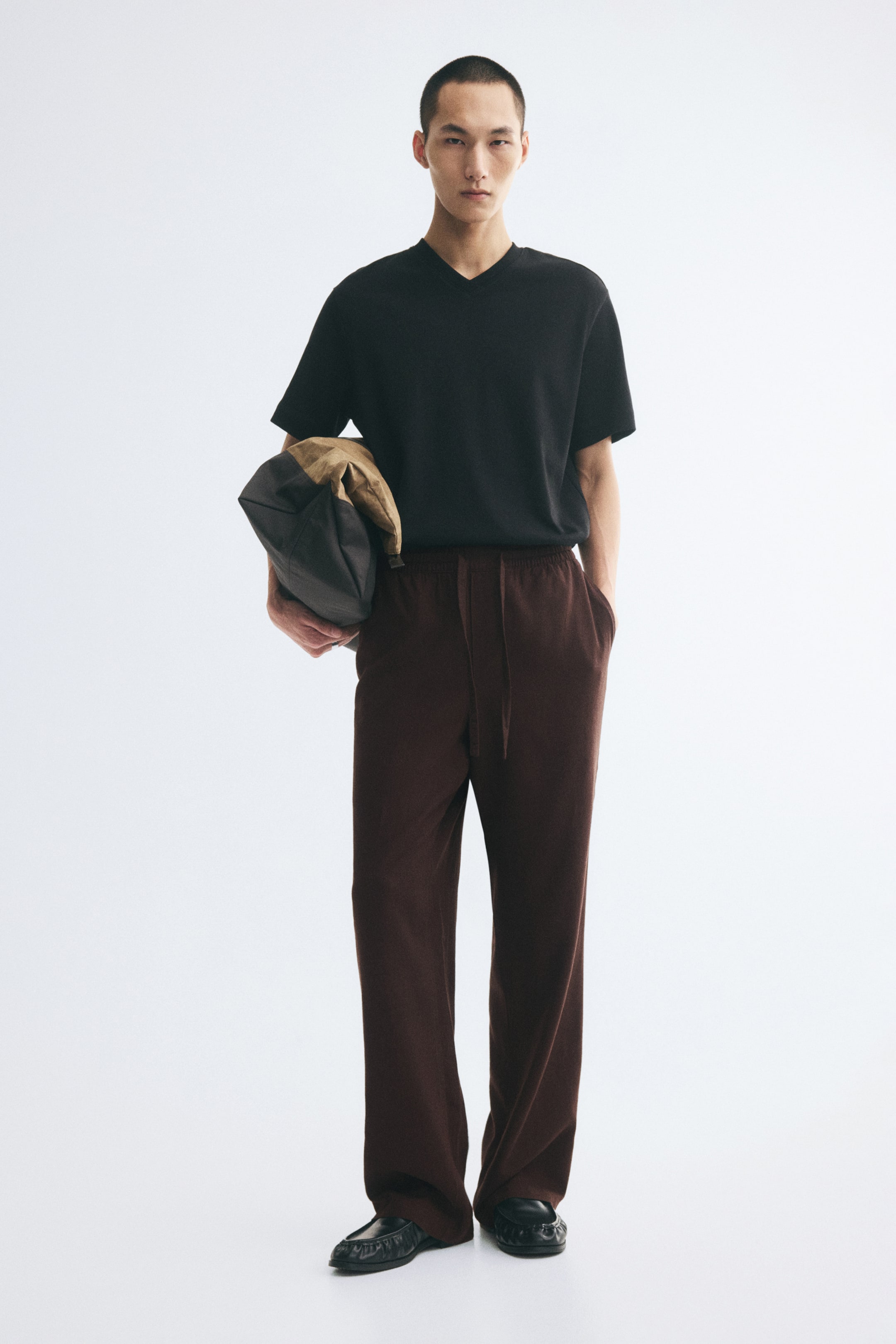 View larger image: Dark brown, relaxed-fit trousers featuring an elasticated drawstring waist and diagonal side pockets are worn by the model, who is standing with one hand in a pocket. The trousers are styled with a tucked-in black V-neck T-shirt, black shoes, and a brown and tan bag carried under one arm.