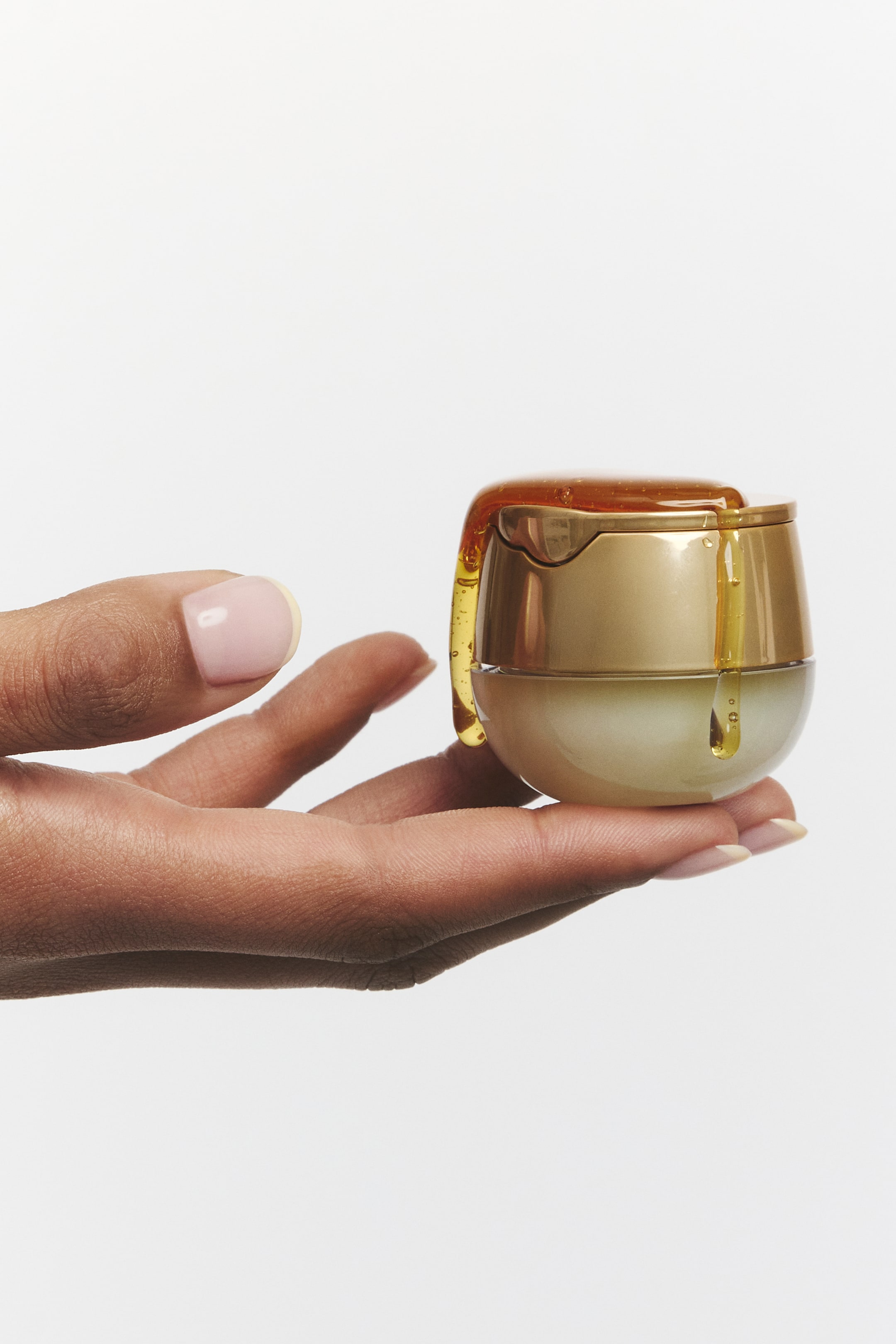Visualizza immagine più grande: Un piccolo vasetto cosmetico arrotondato è rivolto in avanti, tenuto in mano. Presenta un coperchio dorato e una base più chiara, con un liquido simile al miele che gocciola sul coperchio dorato e lungo i suoi lati.