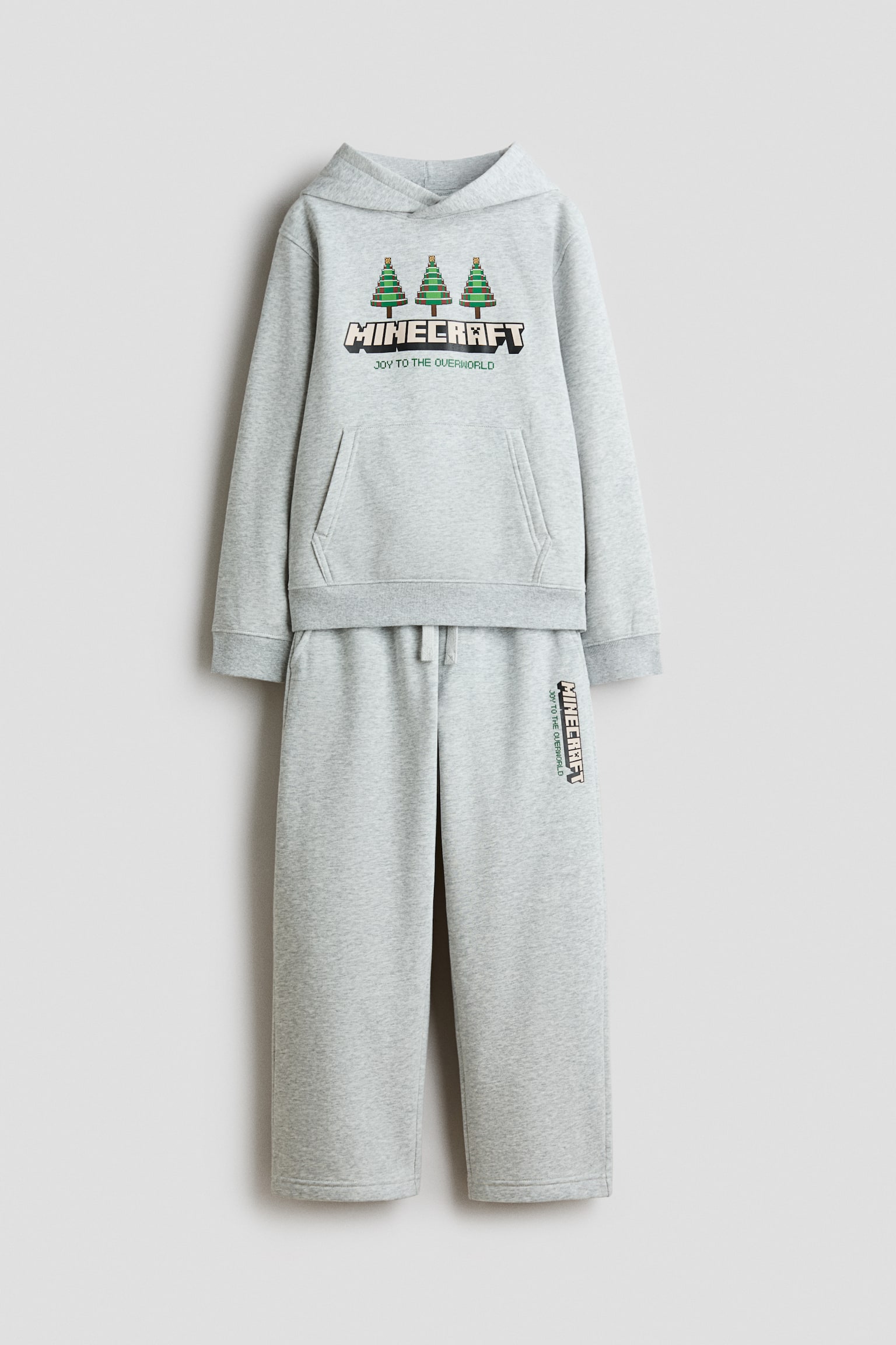 A grey two-piece tracksuit set is shown from the front, featuring a hoodie and matching straight-leg joggers. The hoodie displays three pixelated green trees above a blocky âMINECRAFTâ logo with the text âJOY TO THE OVERWORLDâ, alongside a kangaroo pocket and ribbed cuffs. The matching joggers feature an elastic drawstring waistband and a vertical âMINECRAFTâ graphic printed on the upper leg.