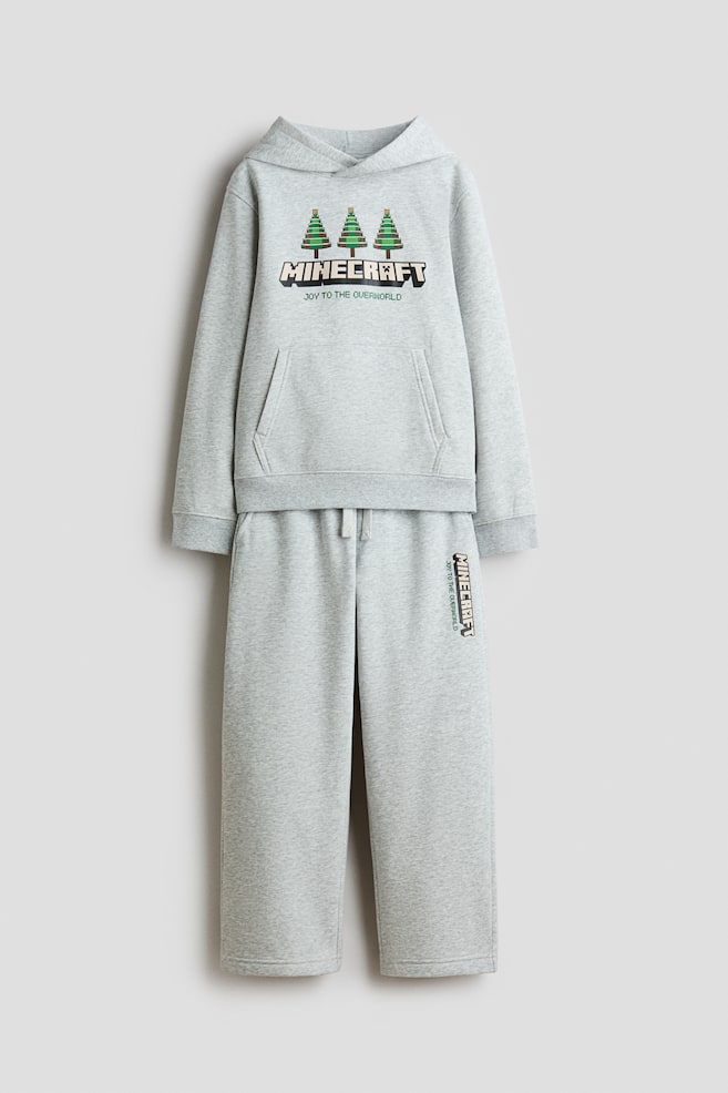 A grey two-piece tracksuit set is shown from the front, featuring a hoodie and matching straight-leg sweatpants. The hoodie is displaying three pixelated green trees above a blocky "MINECRAFT" logo with the text "JOY TO THE OVERWORLD," alongside a kangaroo pocket and ribbed cuffs. The matching sweatpants are featuring an elastic drawstring waistband and a vertical "MINECRAFT" graphic printed on the upper leg. - 1