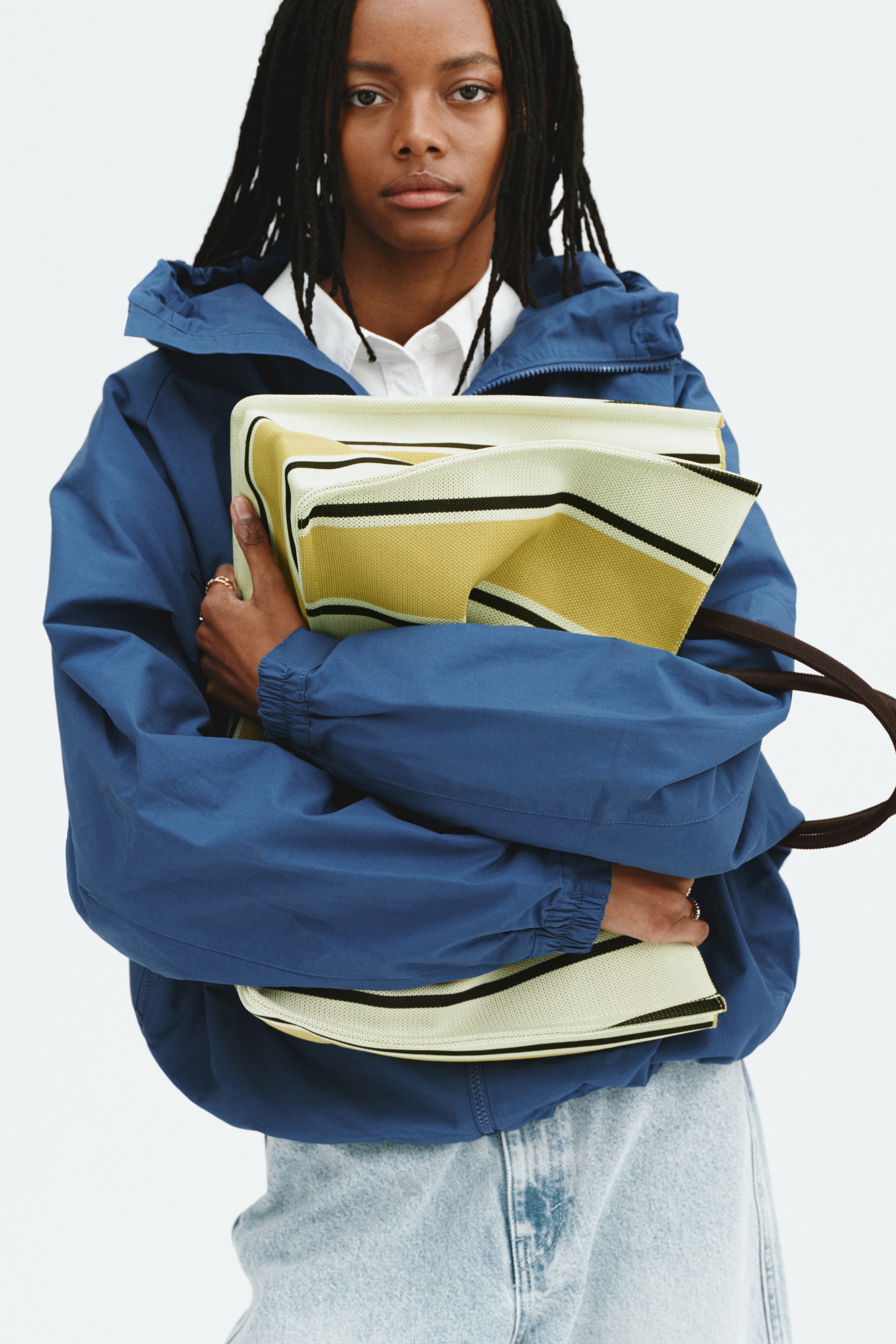 Grösseres Bild ansehen: Die cremefarbene Tasche mit gelb-schwarzen horizontalen Streifen und perforierter Textur wird zusammengefaltet am Körper gehalten, wobei der schwarze Riemen oben sichtbar ist.