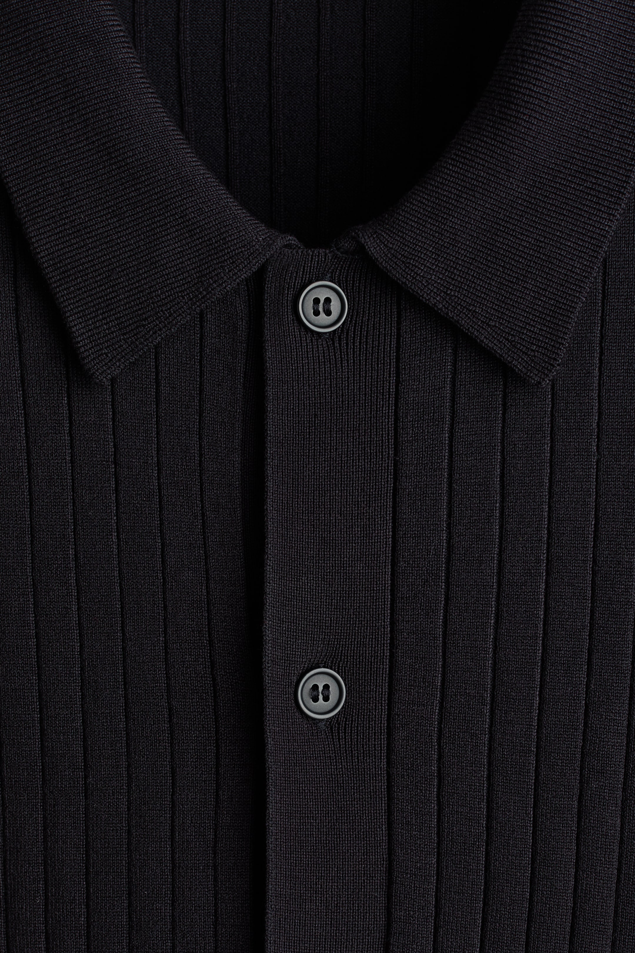 Agrandir l'image: Un polo noir présente une texture en maille côtelée sur le DEVANT. Il est doté d'un col classique et d'une patte de boutonnage avec deux boutons sombres visibles.