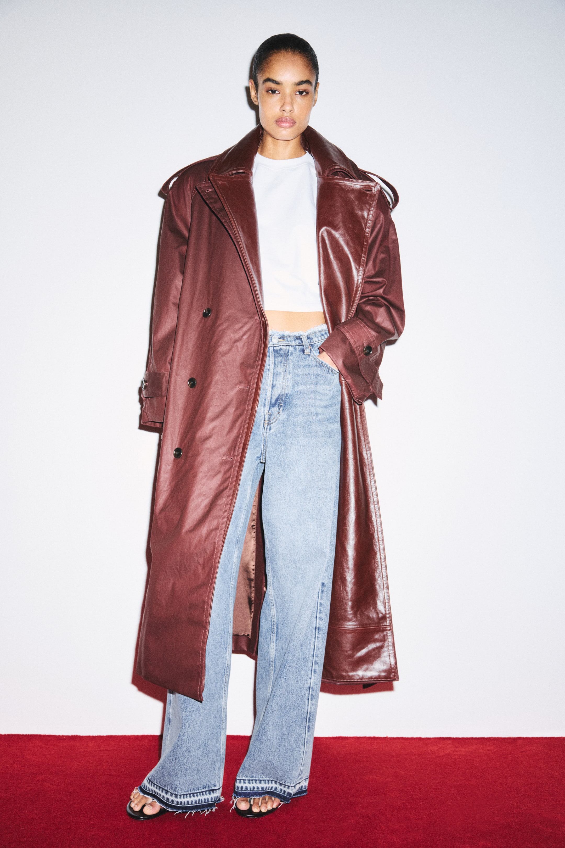 Grotere afbeelding bekijken: Een oversized trenchcoat met dubbele rij knopen in een donker roodbruin gecoate stof wordt open gedragen door het model. De jas heeft brede revers met inkeping en een gekreukelde leren toplaag, epauletten met knopen en een relaxte pasvorm, gecombineerd met een witte cropped T-shirt en lichtblauwe wide-leg jeans.
