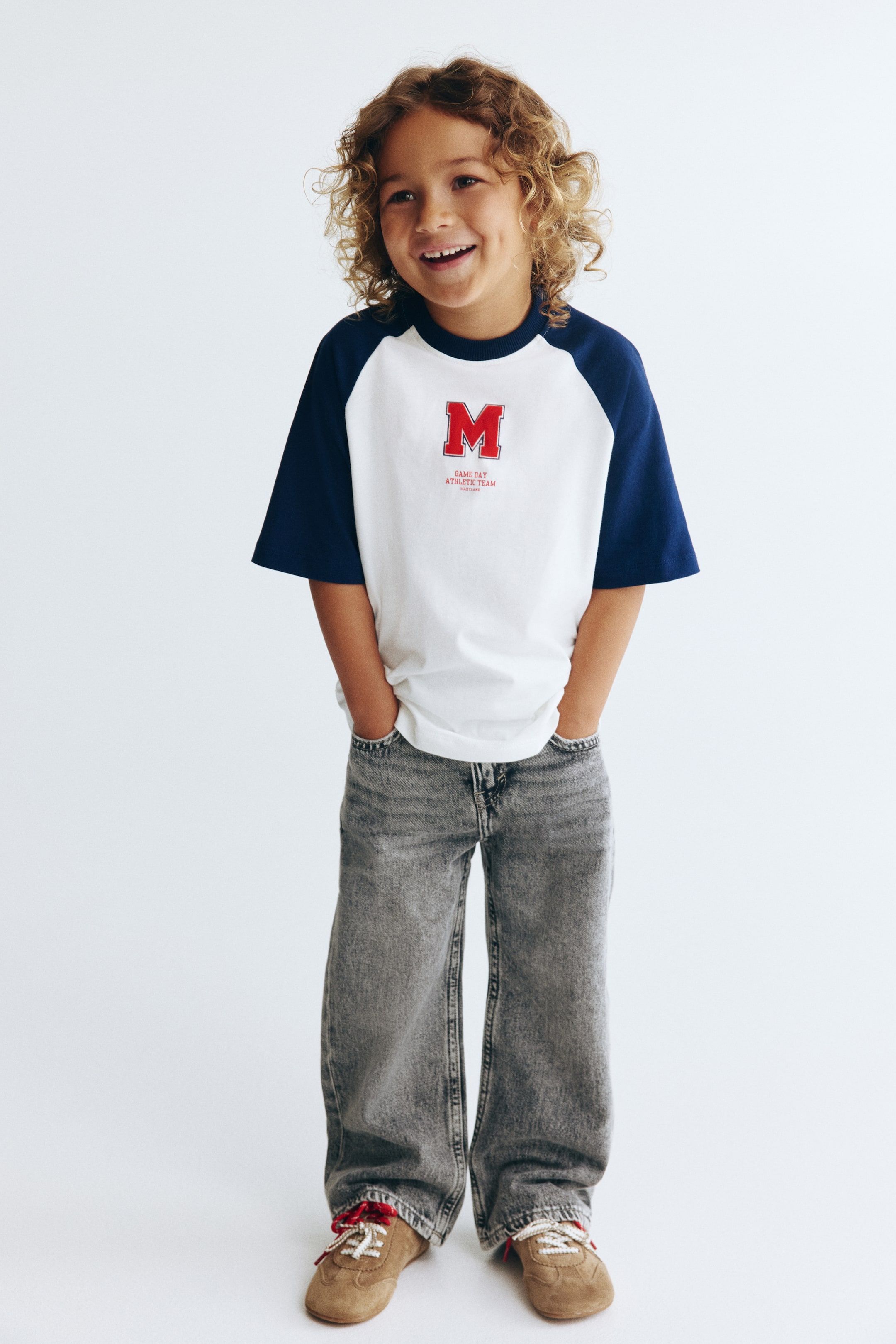 Prikaži veću sliku: Majica sa belom osnovom i tamnoplavim kratkim raglan rukavima, prikazana s prednje strane. Na prednjoj strani nalazi se crveni grafički znak 'M' sa tekstom 'GAME DAY ATHLETIC TEAM', dok okrugli, tamnoplavi rebrasti okovratnik upotpunjuje dizajn.