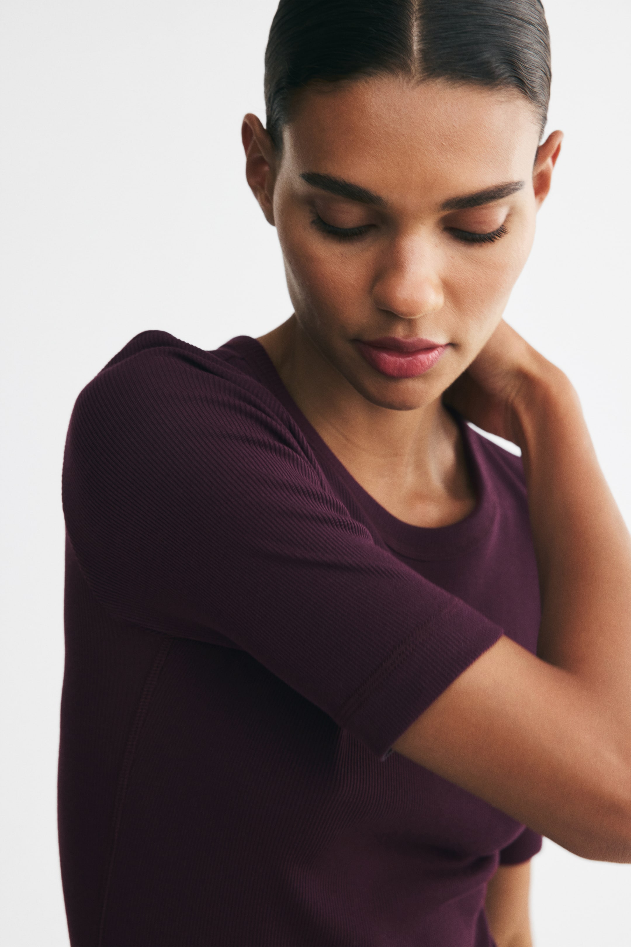 View larger image: A fitted, ribbed jersey top in a dark plum colour, featuring a round neckline, elbow-length sleeves, and visible flatlock seams, is being worn by the model. The model is gently touching her neck, drawing attention to the top's neckline and shoulder.