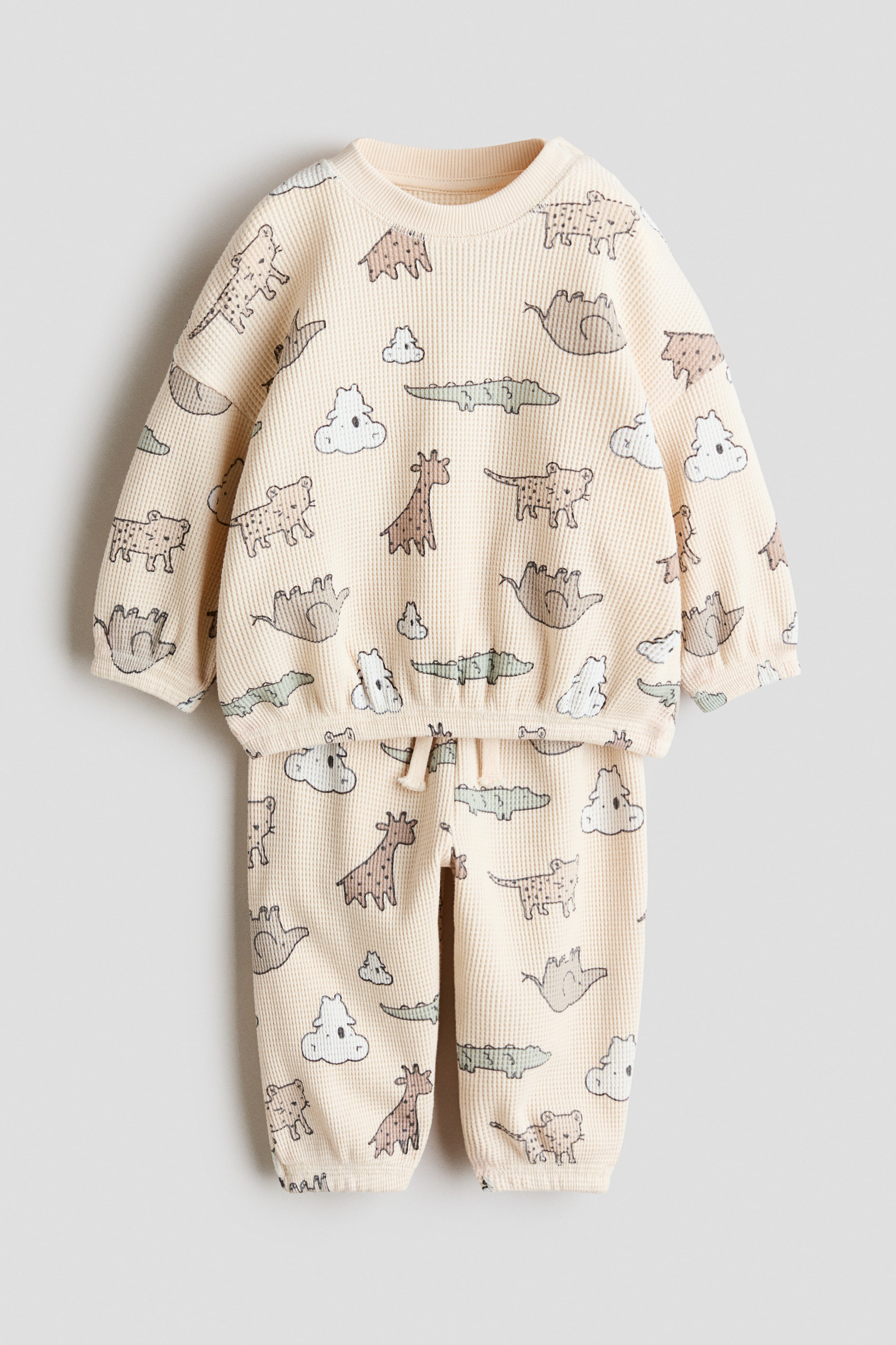 View larger image: A light beige two-piece waffle knit set is shown from the front, comprising a long-sleeved top and matching pants. The set features an all-over print of various cartoon animals and clouds. The top is designed with a crew neck, elasticated cuffs, and an elasticated hem, while the pants include an elasticated drawstring waistband and elasticated cuffs.