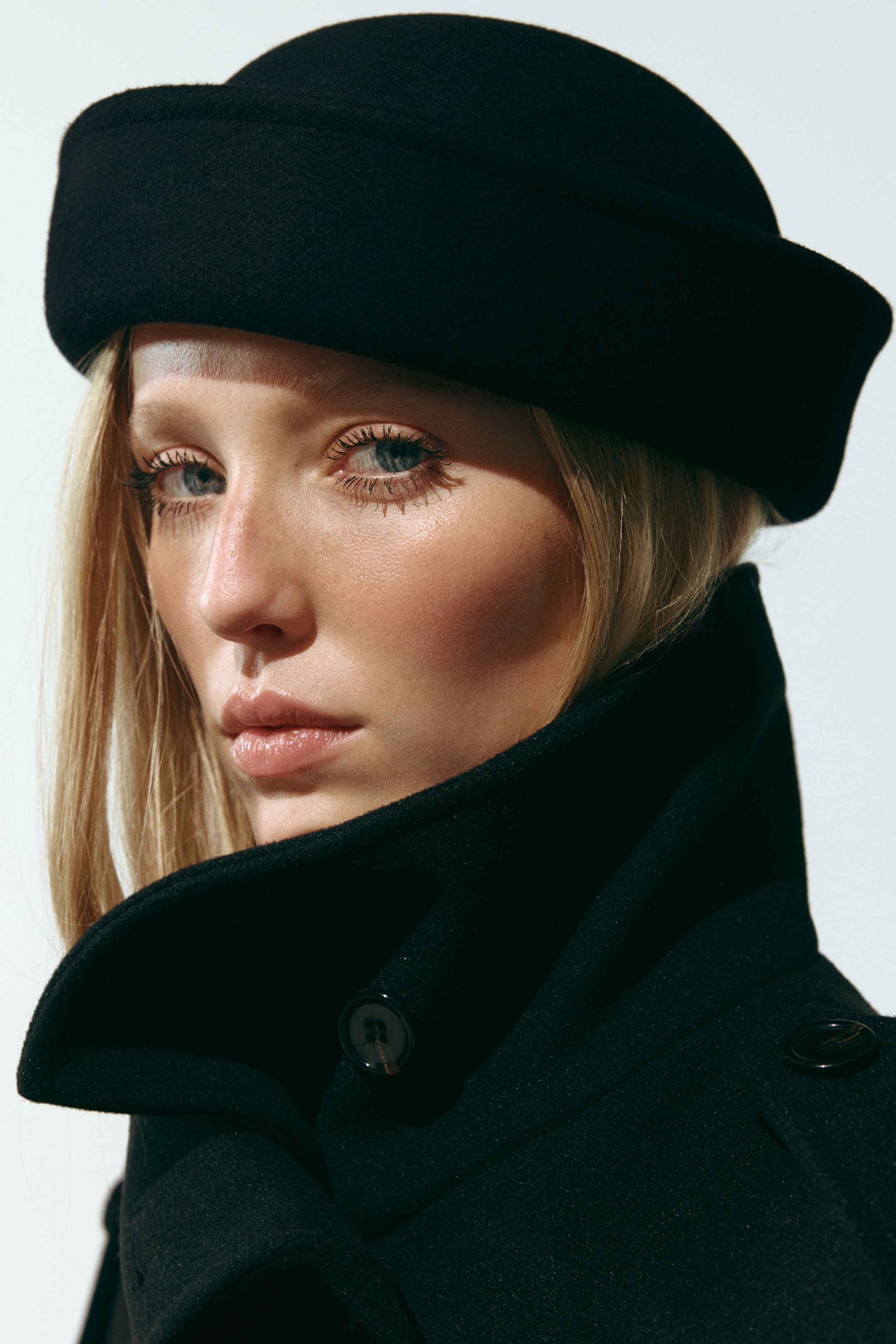 View larger image: A black felted wool hat with a structured, rolled edge is being worn by the model, positioned low on the forehead. The hat is styled with a matching black coat that features a high, turned-up collar.