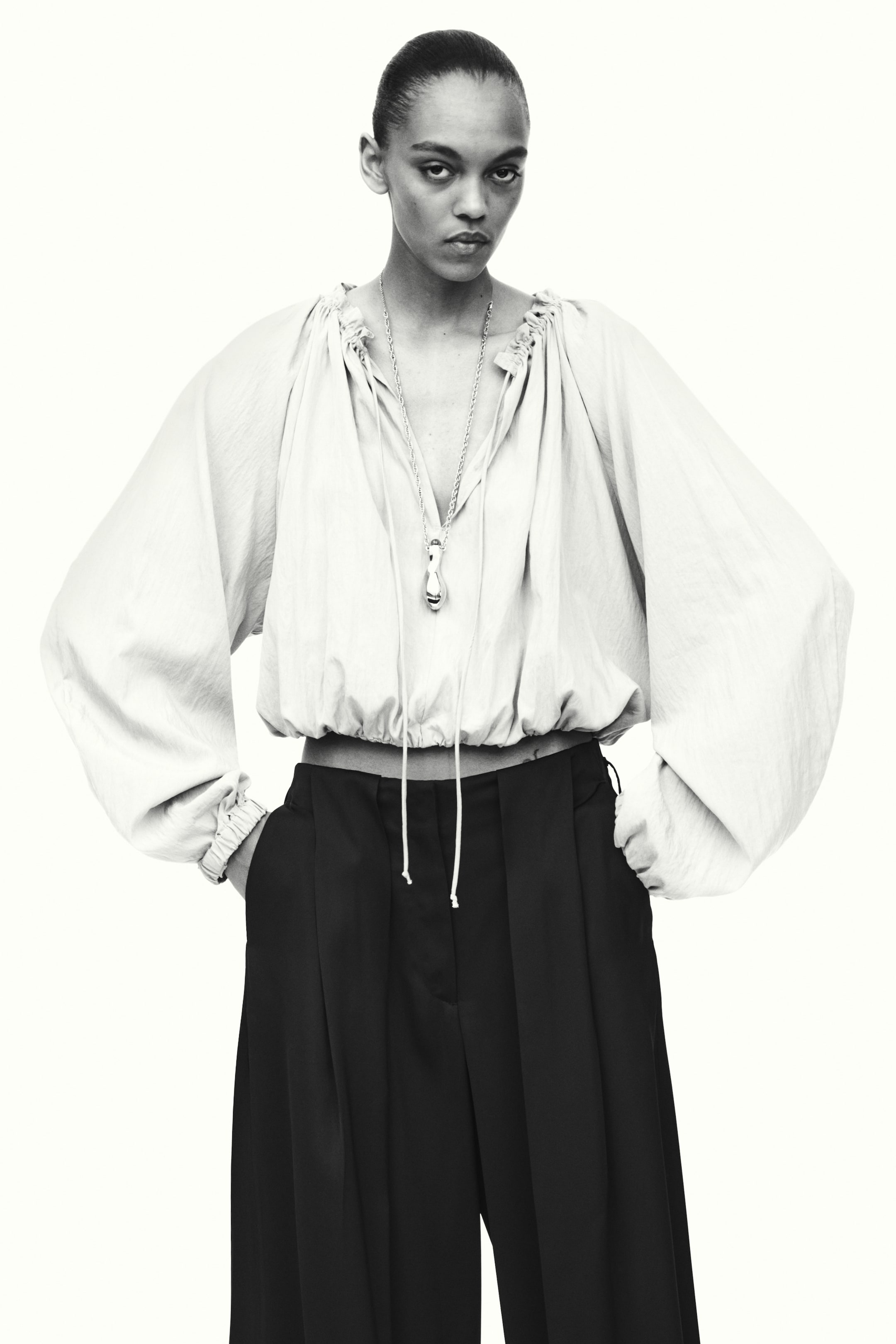 View larger image: A light-colored blouson top features a gathered neckline with visible drawstring ties, voluminous long sleeves with elastic cuffs, and an elastic hem. It's styled over dark, wide-leg trousers with front pleats and a drawstring waist, while a silver-toned necklace with an abstract pendant rests on the top.