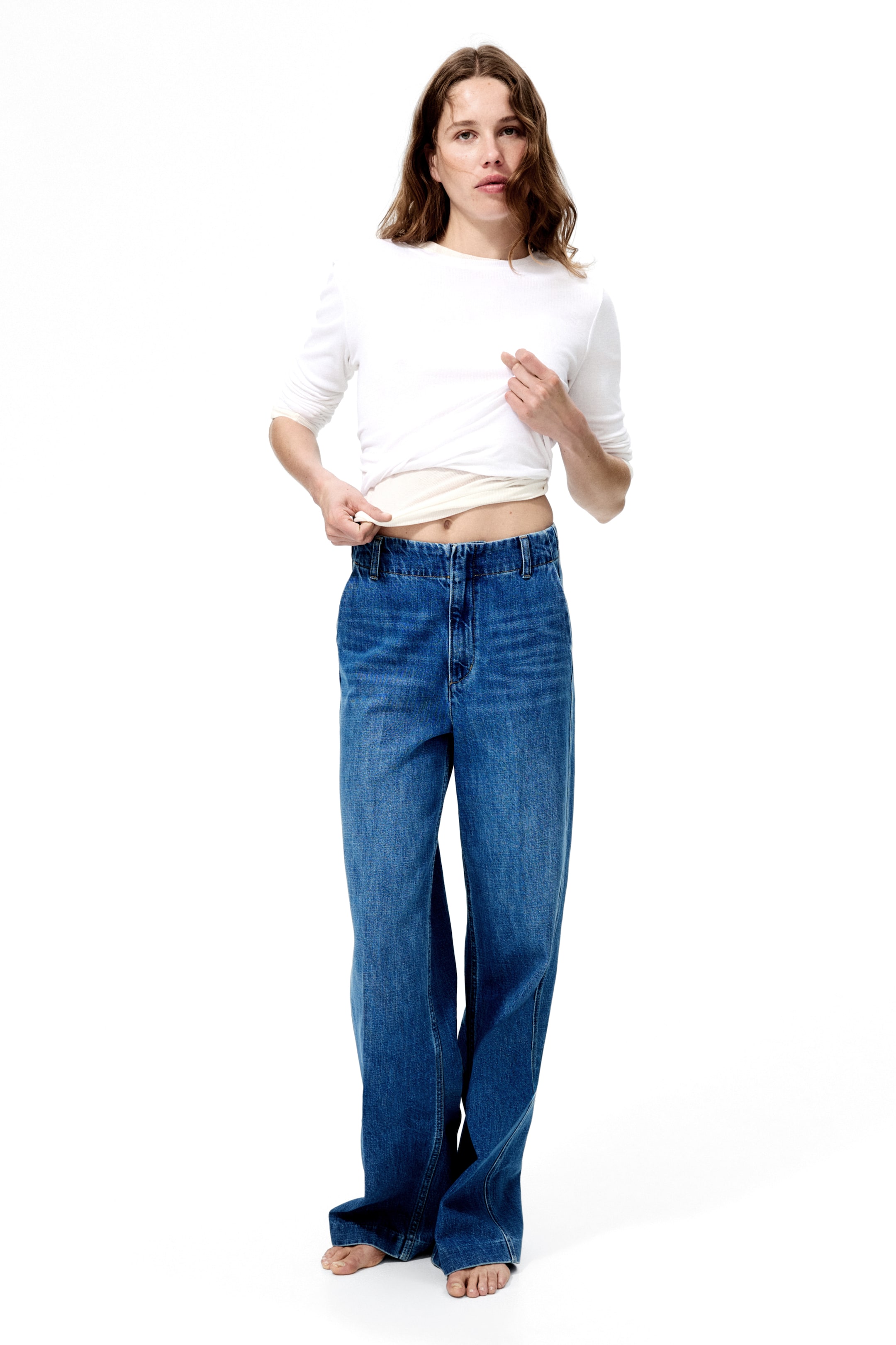 Büyük resmi görüntüle: Mavi geniş paça denim kot pantolonlar, hafif sol ön açıdan sunuluyor, yüksek bel, görünür ön fermuar ve rahat bir kesim gösteriyor. Kumaş ayaklarda toplanıyor.
