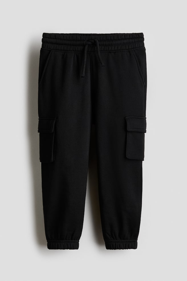 Black cargo sweatpants are featuring an elasticated drawstring waistband, side slash pockets, and flap-closure cargo pockets on the thighs. They are ending with elasticated cuffs at the ankles. - 1