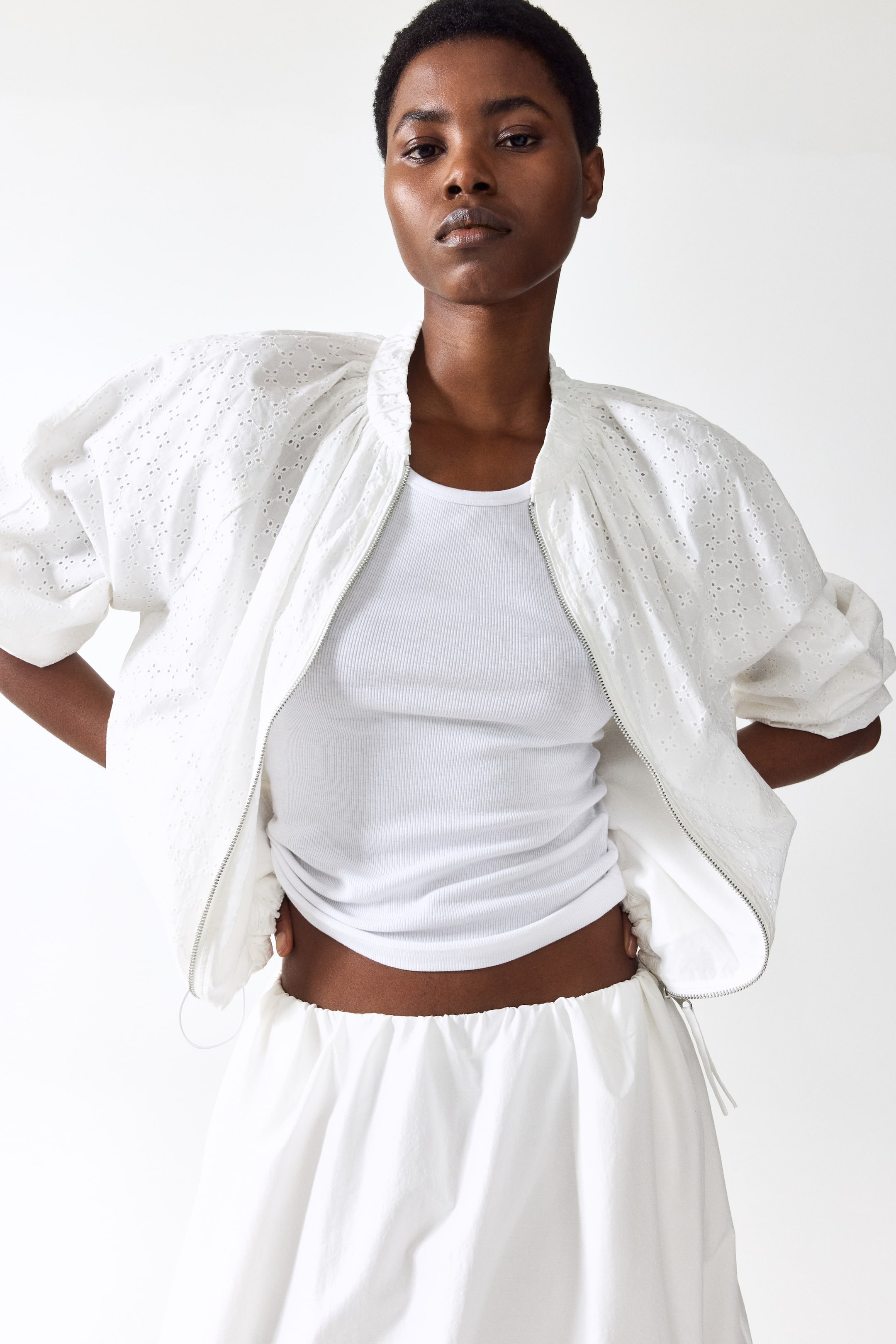View larger image: The model is wearing an oversized white jacket with broderie anglaise detailing. It features a pleated stand collar, raglan sleeves with elasticated cuffs, and an elasticated drawstring hem with cord stoppers. The jacket is unzipped at the front, showcasing a white ribbed top underneath, while the model's hands rest on their hips.
