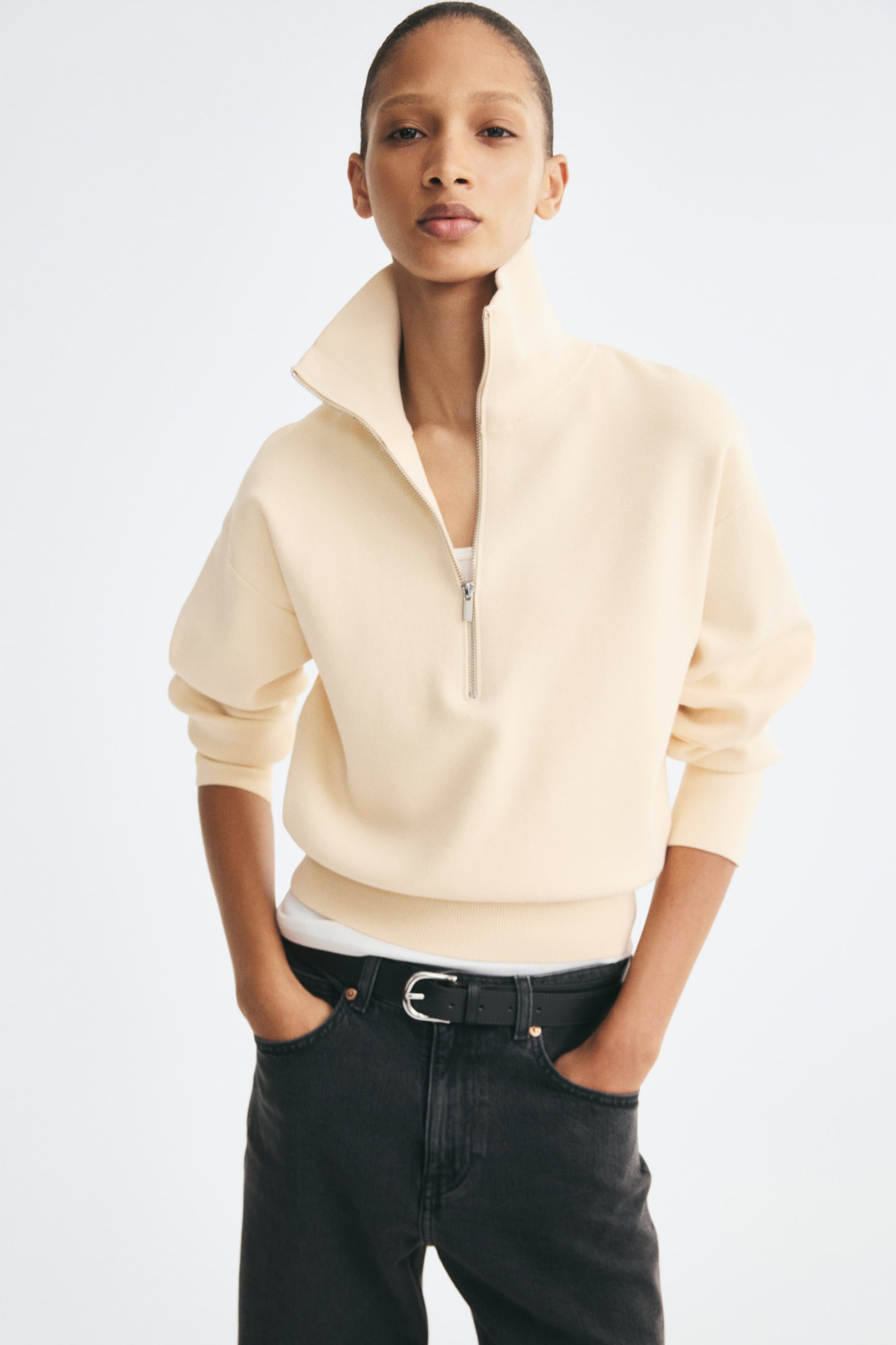 A cream-coloured, fine-knit sweater is shown from the front, featuring a high collar with a partially unzipped half-zip and dropped shoulders. The sweater has long sleeves with ribbed cuffs and a ribbed hem resting above the waistband of black jeans. - 1