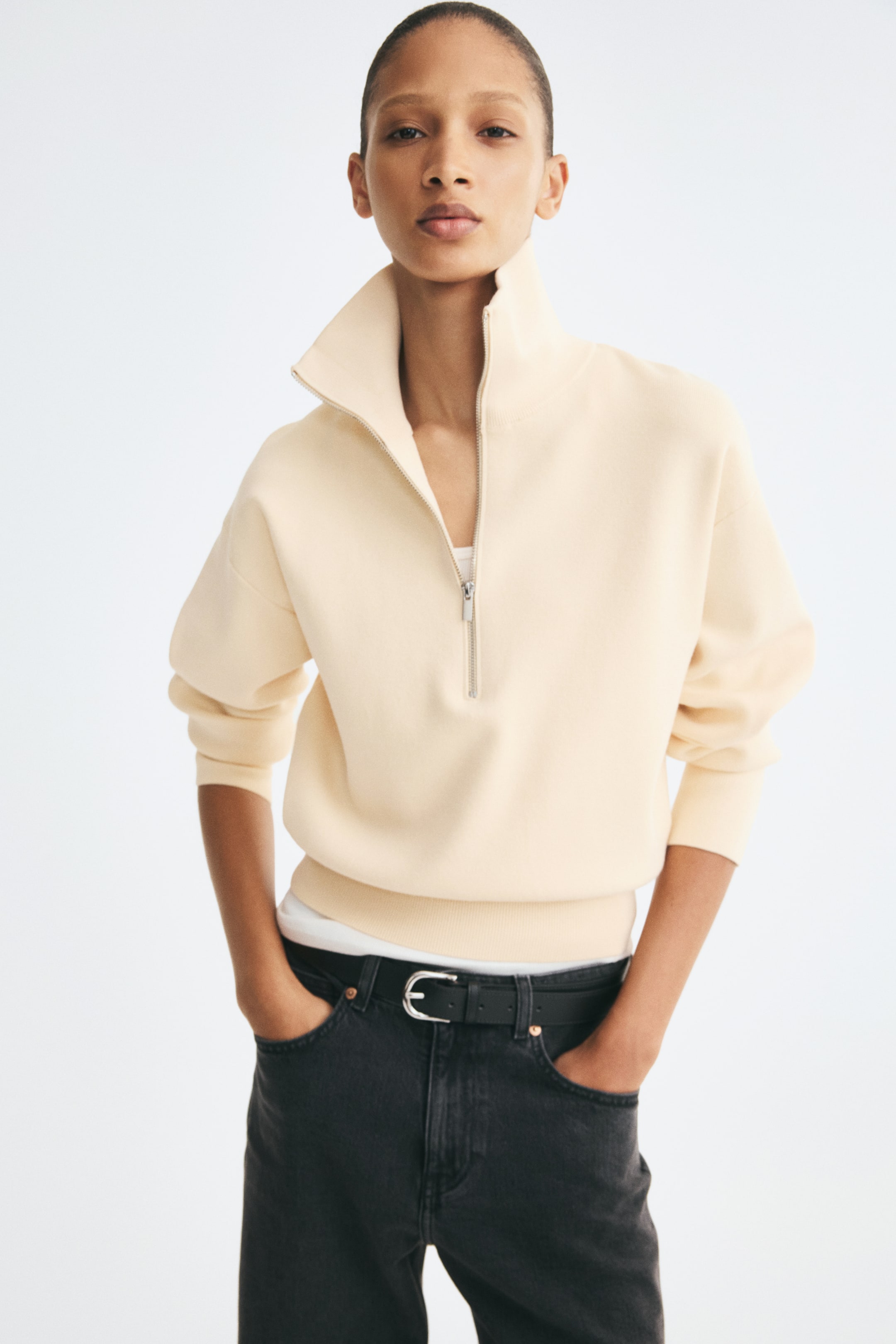 Agrandir l'image: Un pull en maille fine couleur crème est présenté de face, avec un col montant muni d'un demi-zip partiellement ouvert et des épaules tombantes. Le pull présente des manches longues avec des poignets côtelés et un ourlet côtelé arrivant au-dessus de la ceinture d'un jean noir.