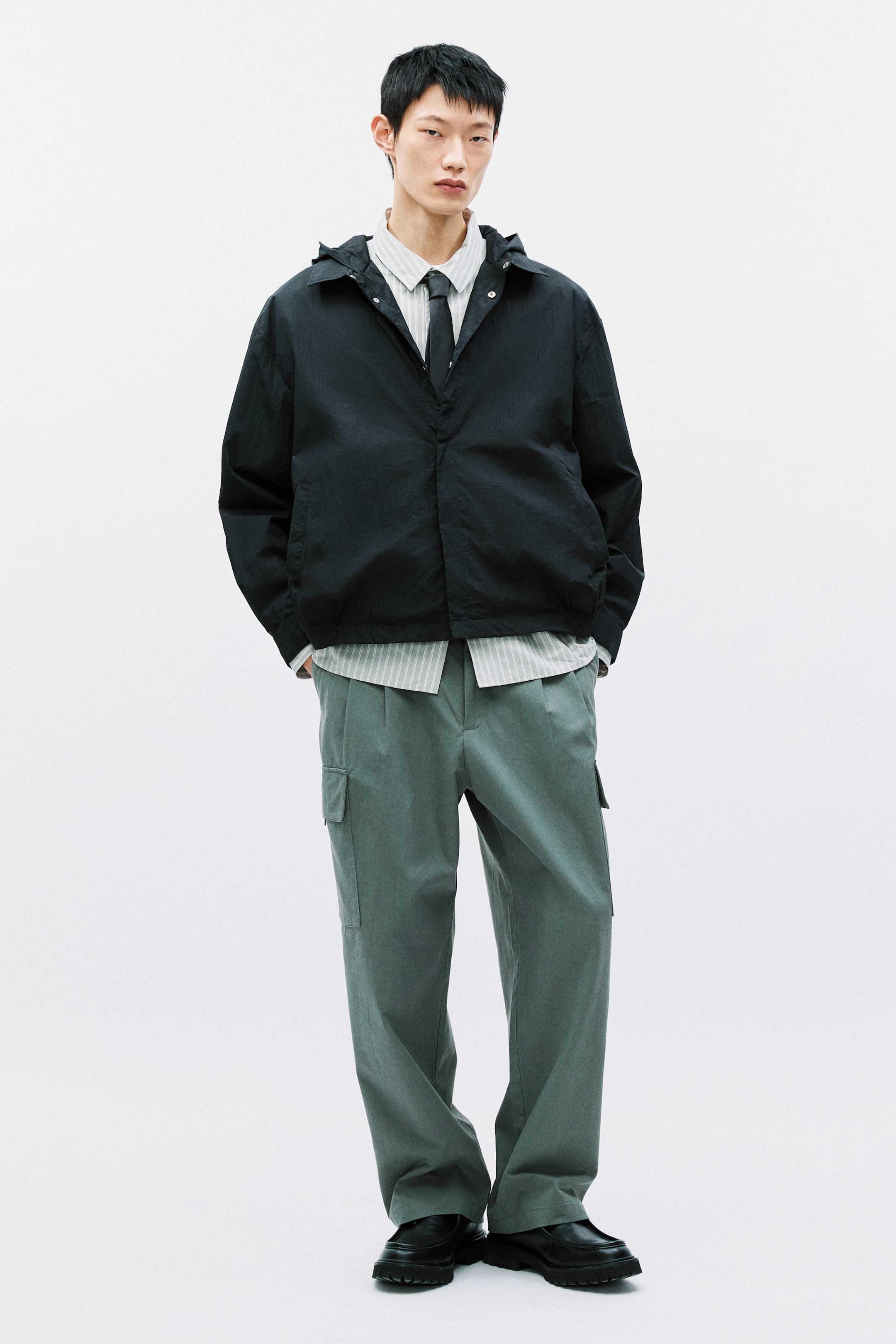 이미지 확대 보기: 어두운 회색 후드 재킷이 소개되고 있으며, 밝은 줄무늬 버튼다운 셔츠와 어울리는 어두운 넥타이 위에 오픈된 상태로 착용되었습니다. 상의와 잘 어울리는 세이지 그린 플리츠 카고 바지는 측면 플랩 포켓이 있으며, 블랙 청키솔 슬립온 슈즈와 함께 착용되었습니다.