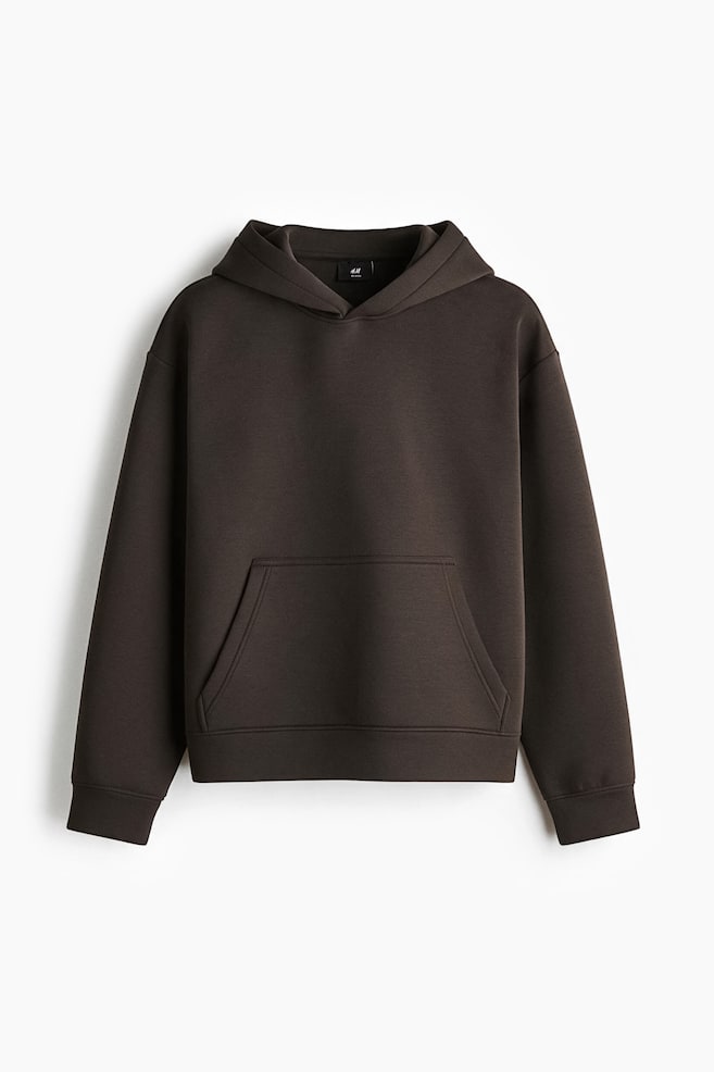 Ein dunkelbrauner Hoodie aus strukturiertem Scuba-Material ist von vorn zu sehen und zeichnet sich durch eine lockere Passform mit Ã¼berschnittenen Schultern und langen Ãrmeln aus. Das Design umfasst eine doppellagige Kapuze, eine KÃ¤ngurutasche auf der Vorderseite sowie breite BÃ¼ndchenabschlÃ¼sse an den Ãrmeln und am Saum. - 1