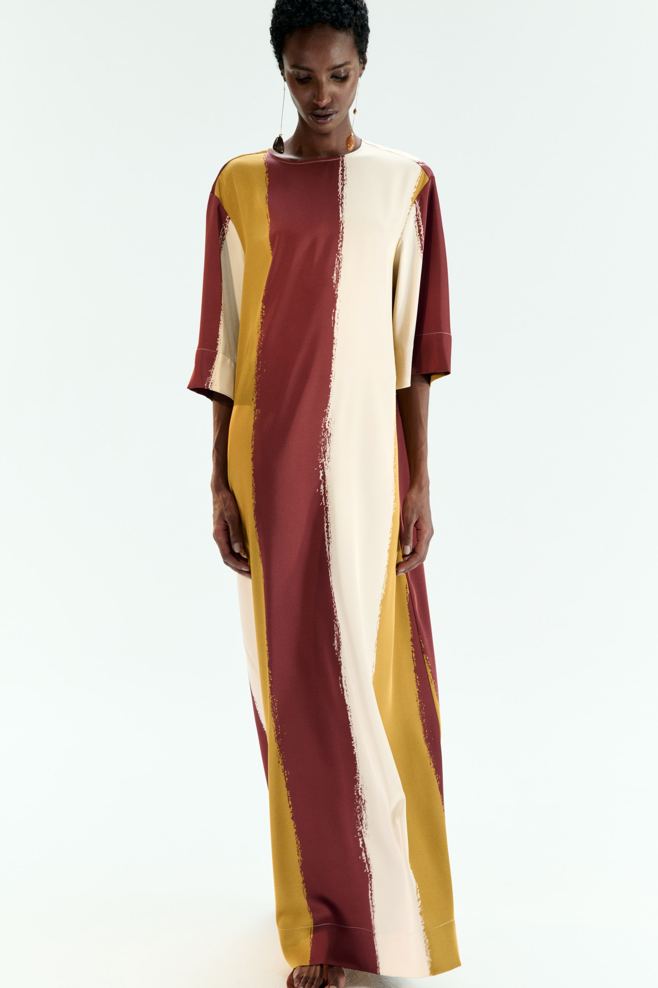 View larger image: A long, loose-fitting dress is displaying wide, vertical brushstroke stripes in cream, golden yellow, and reddish-brown. It is featuring a round neckline and elbow-length sleeves, extending to the floor.