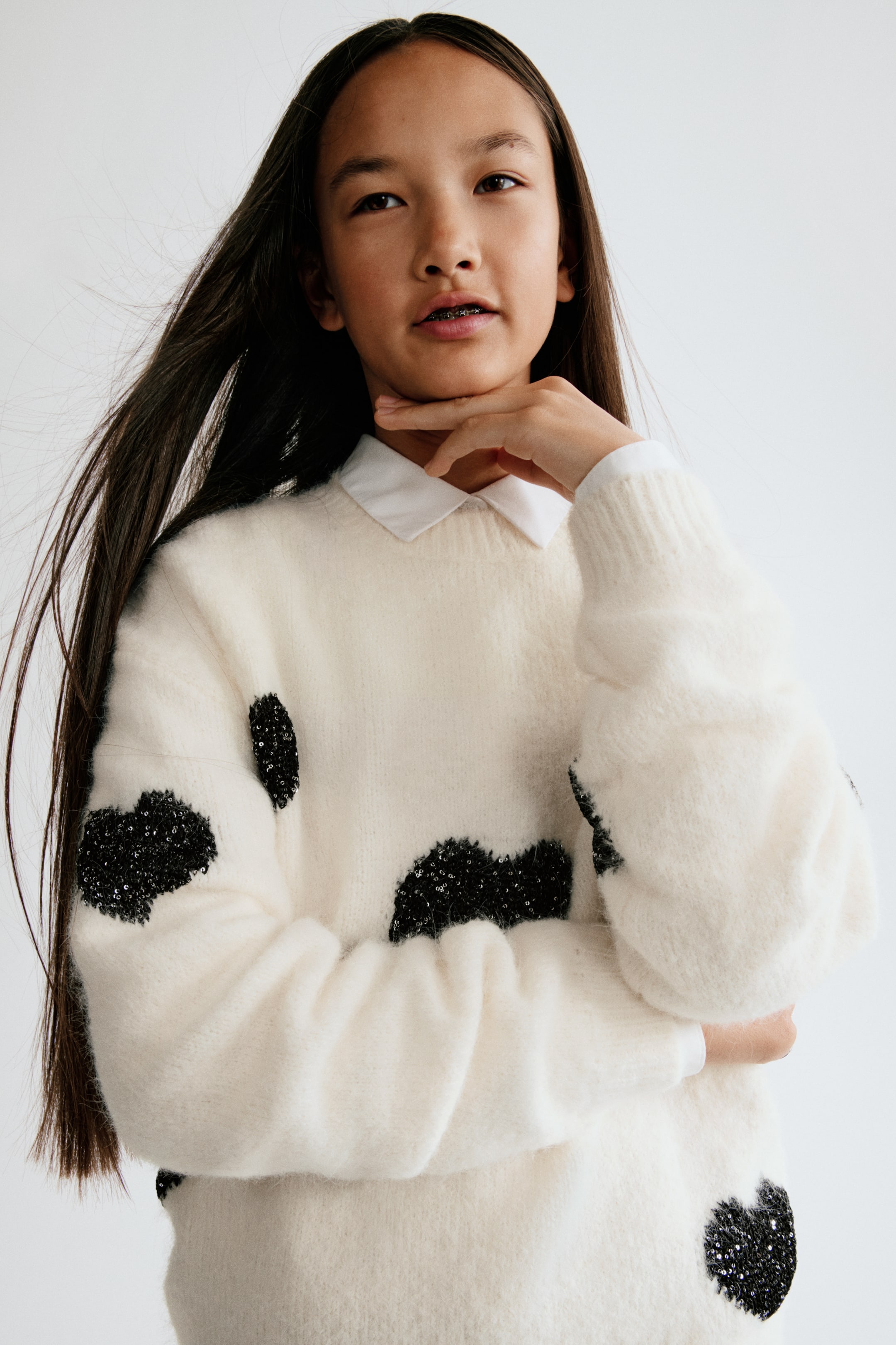 Agrandir l'image: Un pull en maille blanche, au toucher doux et brossé, orné de patches noirs pailletés, présente un col rond en bord-côte, des épaules tombantes et des poignets côtelés. Le pull est présenté de face, superposé à une chemise blanche à col.