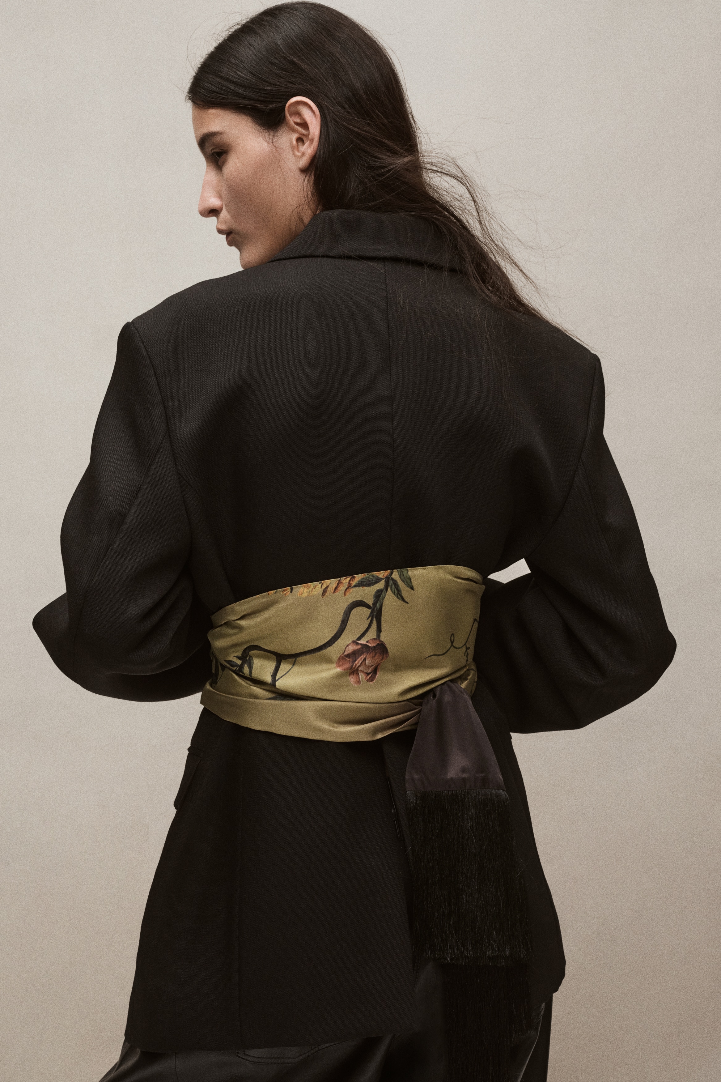 An olive-green satin scarf is draped vertically, displaying a botanical print of pink blossoms, yellow wisteria-like flowers and green foliage. The scarf features a contrasting dark brown underside and finishes with a long, dense black fringe.