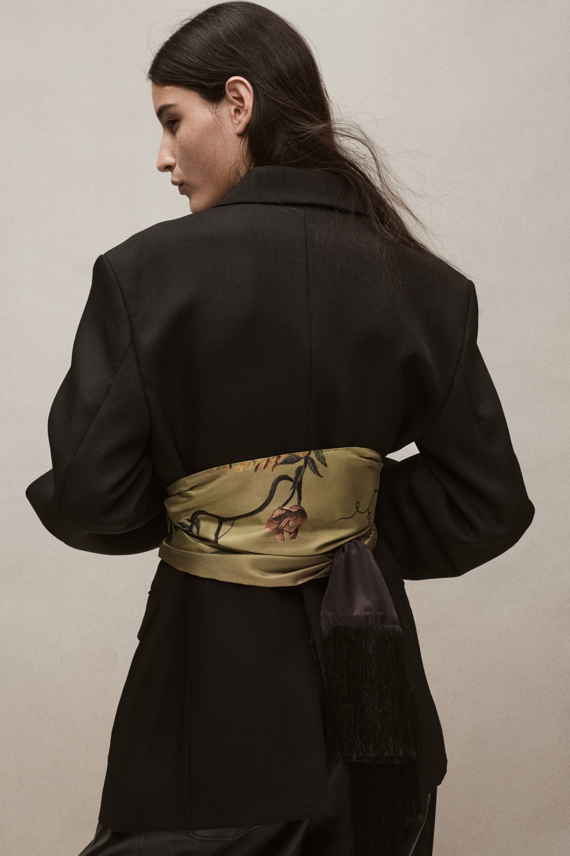View larger image: A satin scarf with a green floral print is styled wrapped around the waist of a black blazer. The scarf is tied in back, with its long black fringed end hanging down.