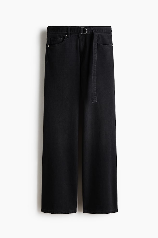 Los pantalones de mezclilla negros de pierna ancha se muestran en vista frontal, con un cinturón de tela negra que cuenta con una hebilla en forma de D de color plateado. El cinturón está pasado por las trabillas y se extiende hacia abajo en la parte delantera. - 1