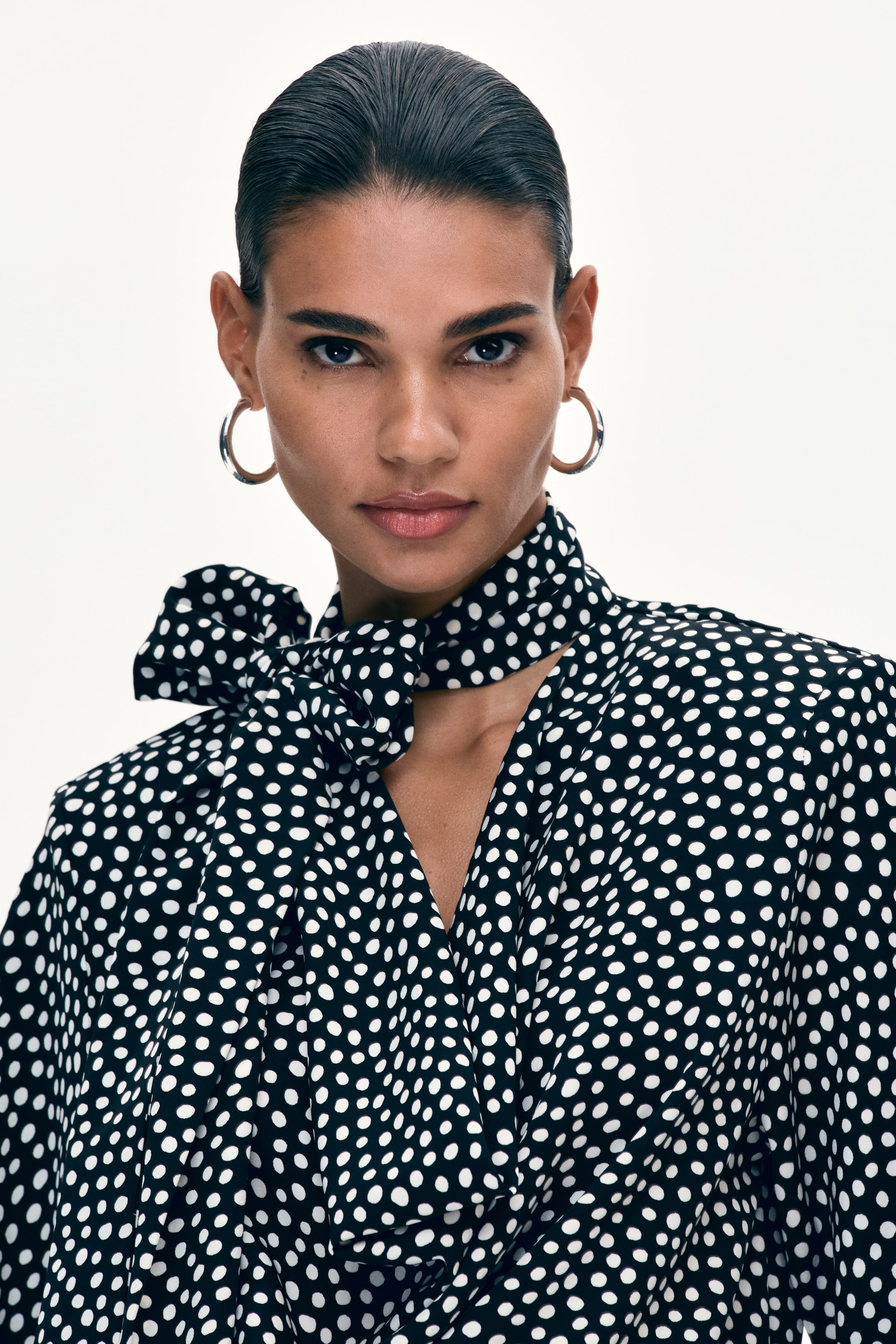 View larger image: A black blouse with scattered white polka dots features a self-tie neck scarf tied into a bow on the left side and a draped V-neckline, as viewed from the front.