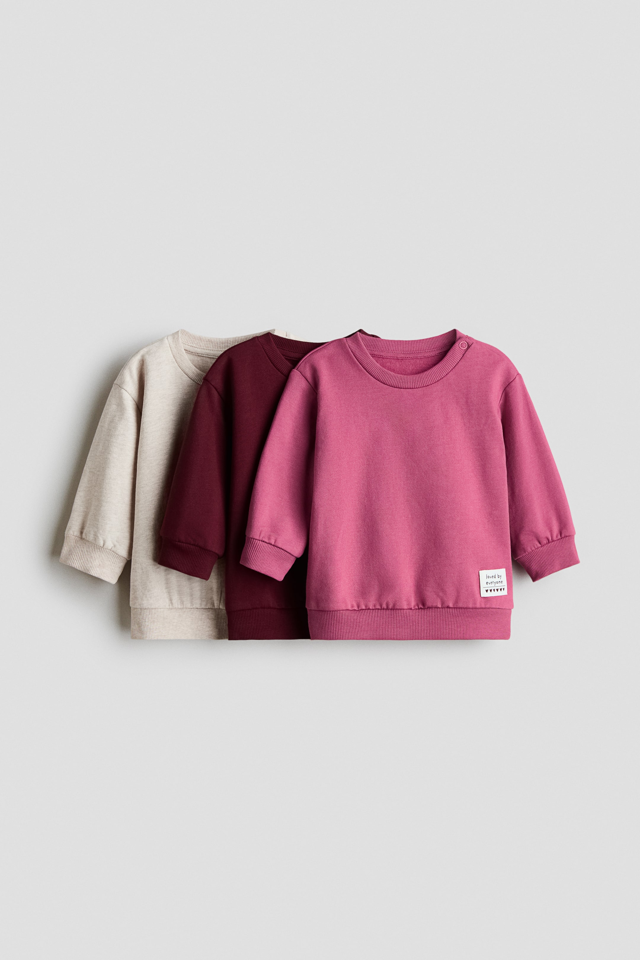 View larger image: A fuchsia baby sweatshirt is shown in the foreground, featuring a round neck, long sleeves with ribbed cuffs, a ribbed hem, and two snap buttons on the left shoulder, along with a small white fabric label on the lower right front. It is layered with a maroon sweatshirt behind it and a beige sweatshirt behind the maroon one, all facing forward at an angle.