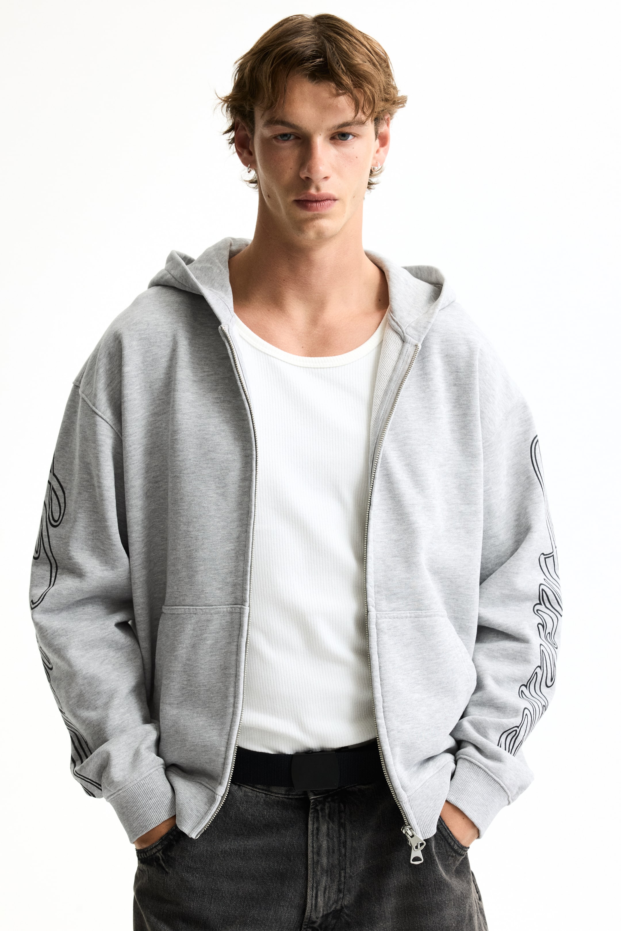 View larger image: A gray zip-up hoodie is showcased with an open, full-length silver-toned zipper, an oversized fit, and black wavy, flame-like graphics adorning the outer sides of both long sleeves. The sleeves conclude with ribbed cuffs, complementing the ribbed hem, and the hoodie is layered over a white ribbed t-shirt.