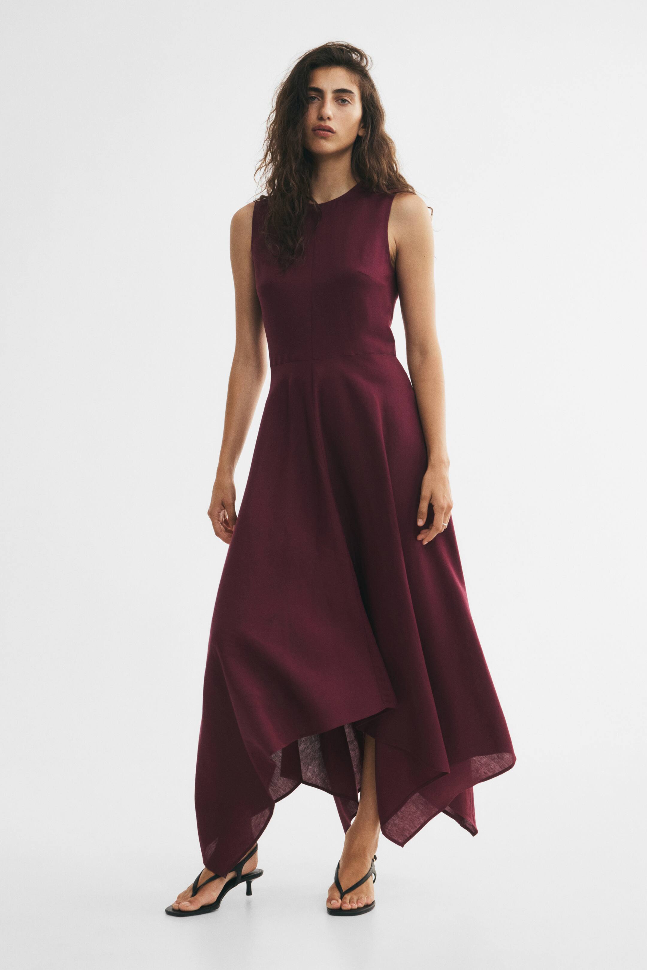 View larger image: A deep red, ankle-length, sleeveless dress is worn by the model, featuring a fitted bodice with a round neckline and a flared skirt with a handkerchief hem. The model is standing and wearing black heeled sandals.