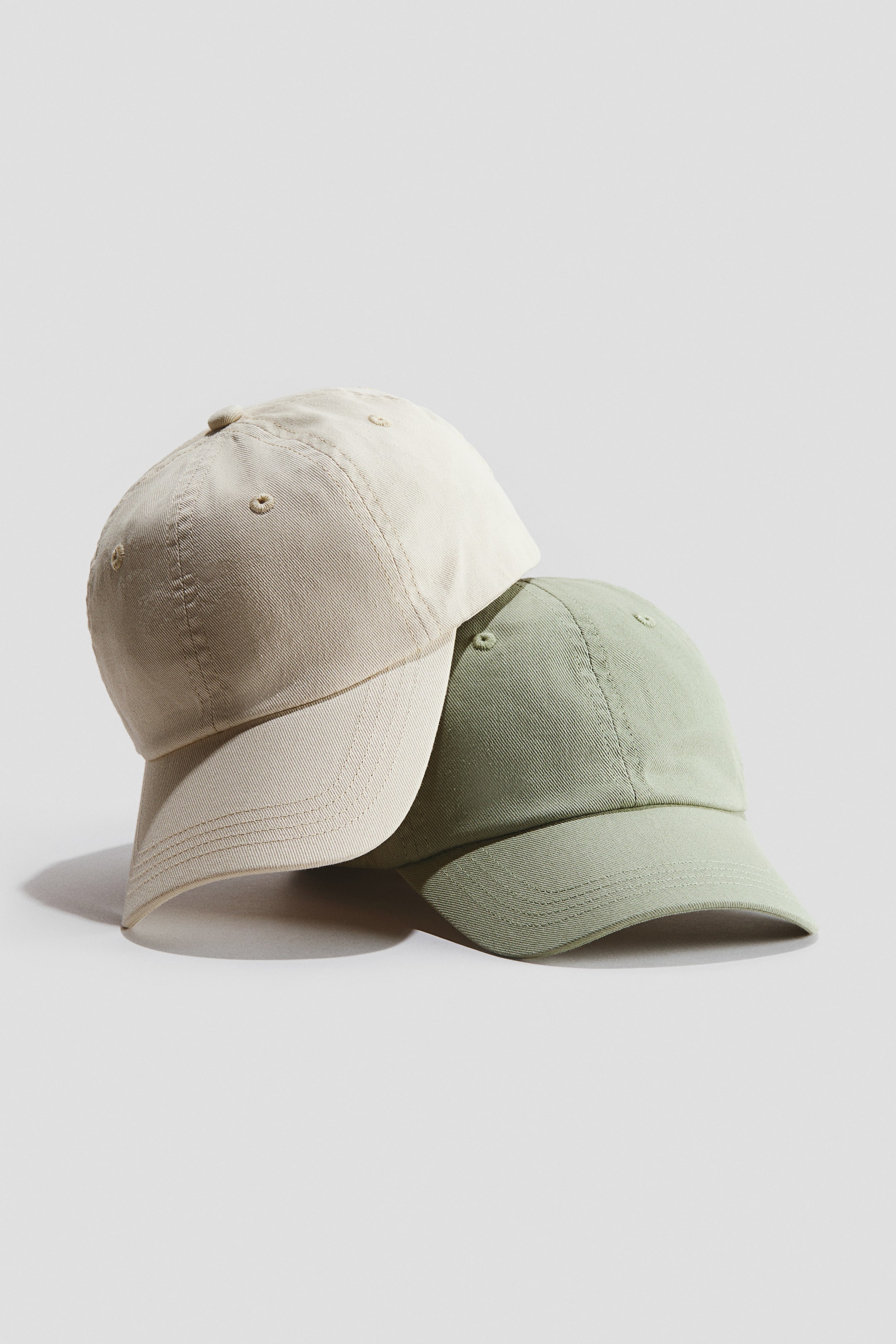 Visualizza immagine più grande: Due cappellini in twill di cotone sono esposti, con le angolazioni frontali e laterali visibili. Un cappellino beige chiaro è posizionato leggermente dietro e sopra un cappellino verde chiaro, entrambi caratterizzati da occhielli ricamati e visiere cucite.