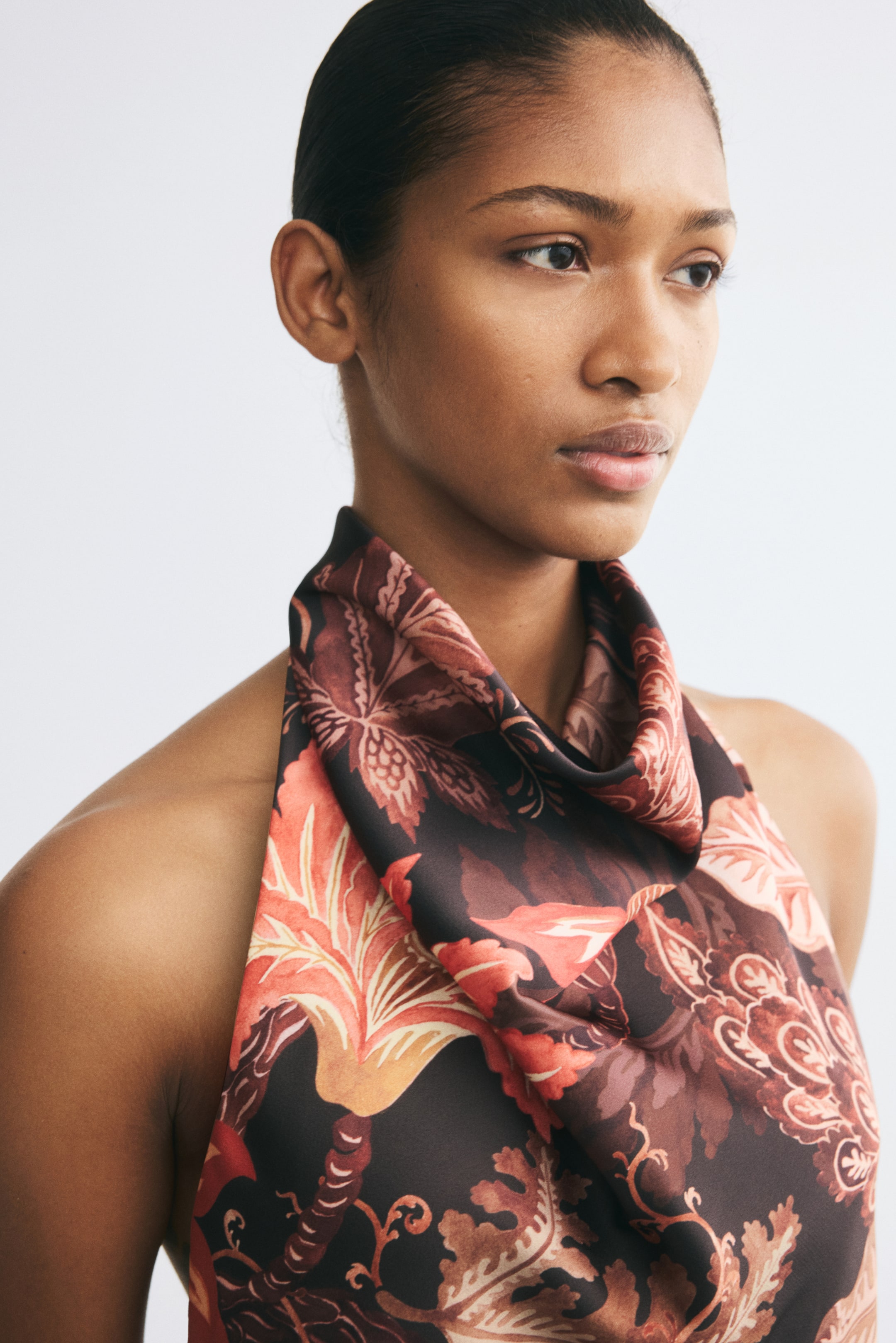 View larger image: The model is wearing a halterneck top with a floral pattern in shades of brown, orange, and pink. The top features a draped cowl neckline and a wide strap that wraps around the neck.