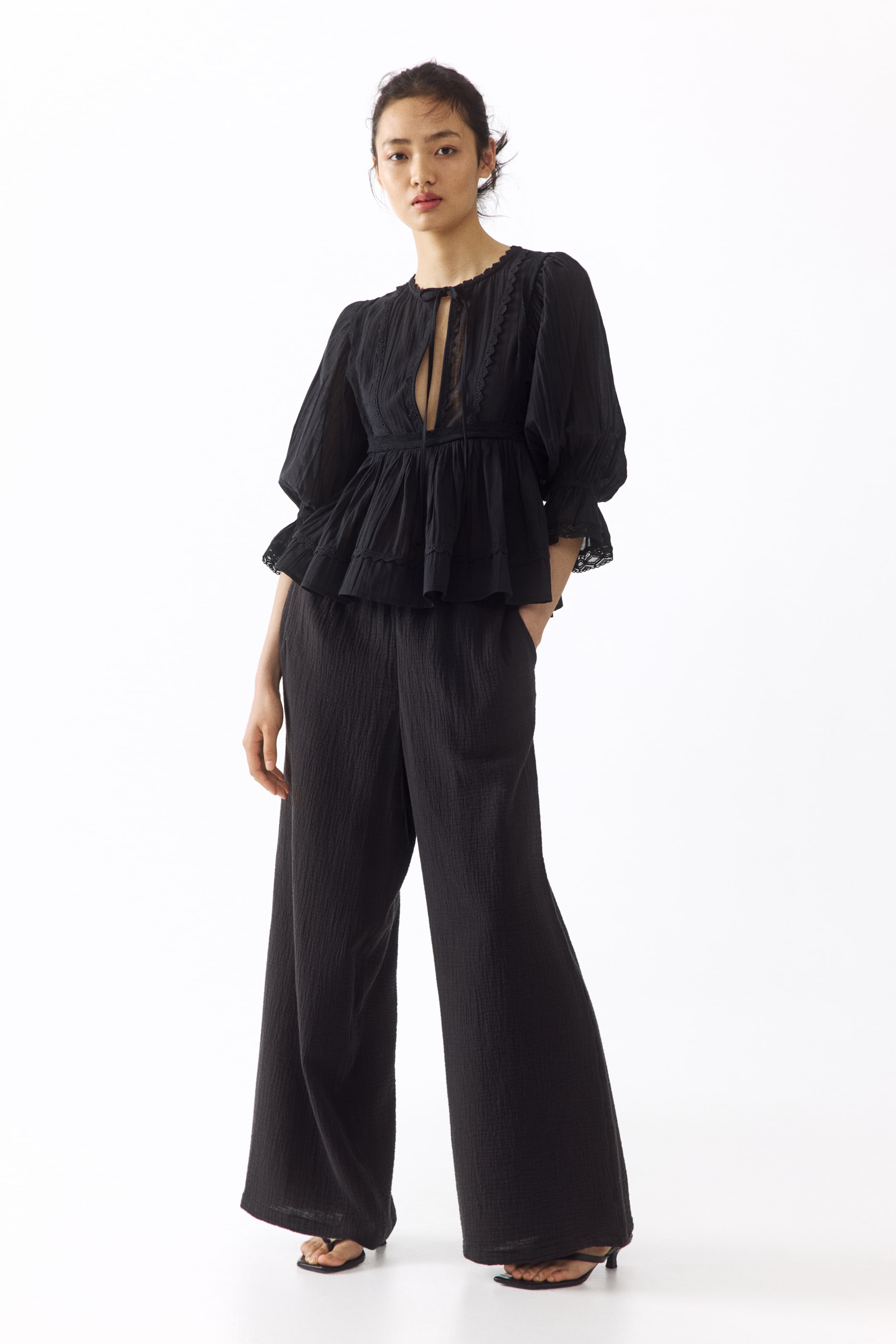 View larger image: A black textured peplum top featuring a round neckline with a keyhole opening and tie closure, scalloped trim, pintucks, lace inserts, and puff sleeves ending in elasticated cuffs with lace trim. The matching black textured wide-leg pants feature side pockets and a full-length silhouette.