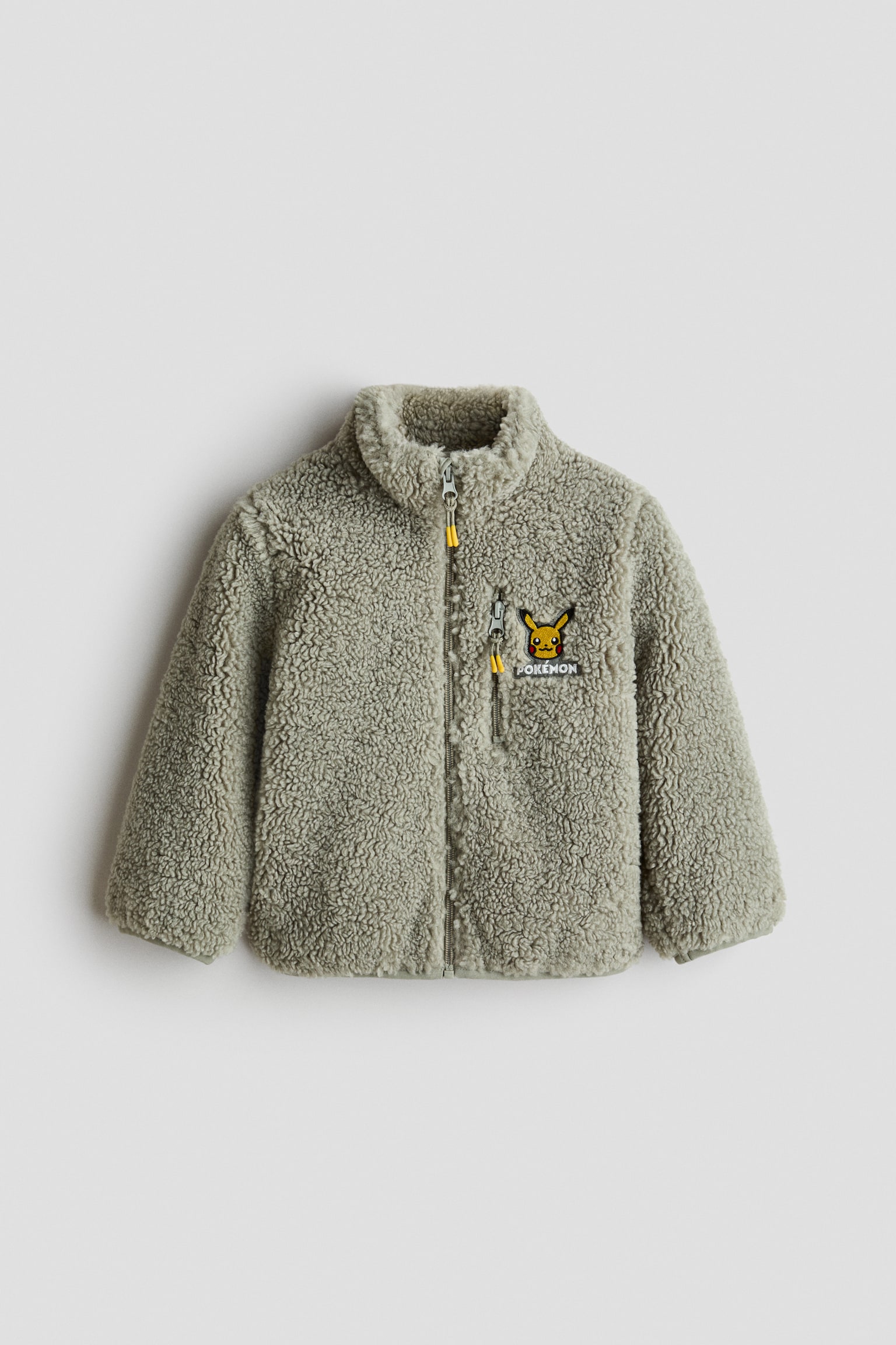 A light green sherpa fleece jacket is shown from the front, featuring a high collar, a full-length zipper with a yellow pull tab, and a vertical zippered chest pocket. An embroidered Pikachu patch with the word "POKï¿½MON" decorates the left chest.