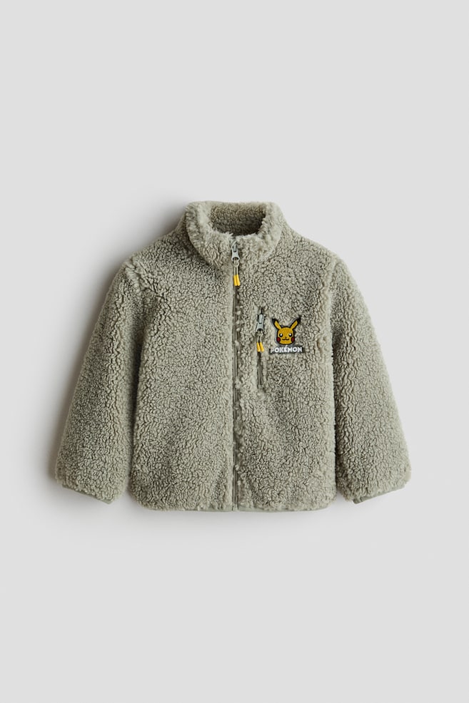 A light green sherpa fleece jacket is shown from the front, featuring a high collar, a full-length zipper with a yellow pull tab, and a vertical zippered chest pocket. An embroidered Pikachu patch with the word "POKÃMON" decorates the left chest. - 1