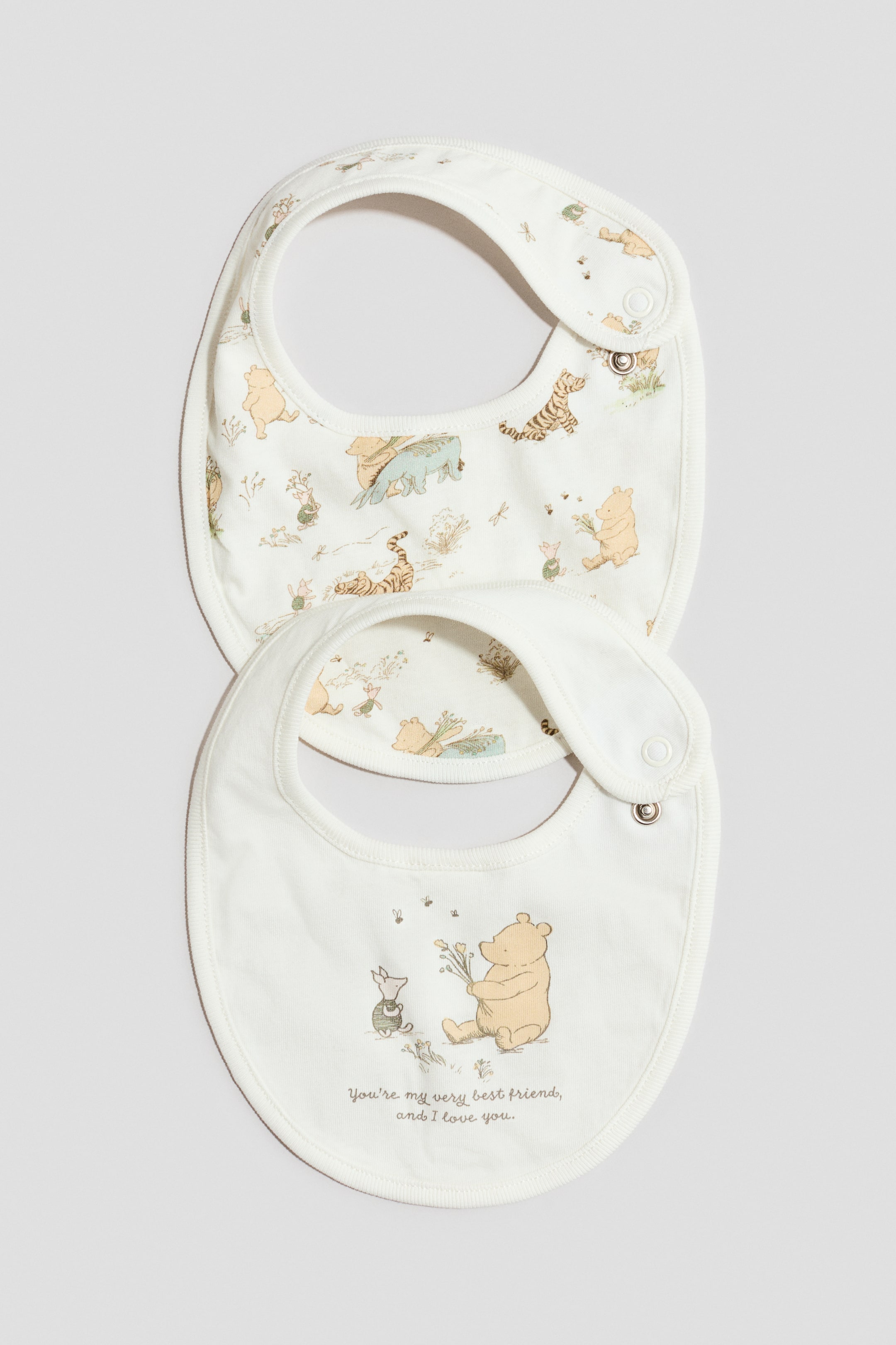 Agrandir l'image: Deux bavoirs pour bébé de couleur crème sont présentés de face, avec des formes arrondies, des bords passepoilés et des fermetures à pression sur le côté. Le bavoir du haut arbore un imprimé intégral classique du personnage Winnie l’Ourson, tandis que celui du bas met en avant un visuel central de Winnie l’Ourson et Porcinet accompagné du texte « You’re my very best friend, and I love you. »