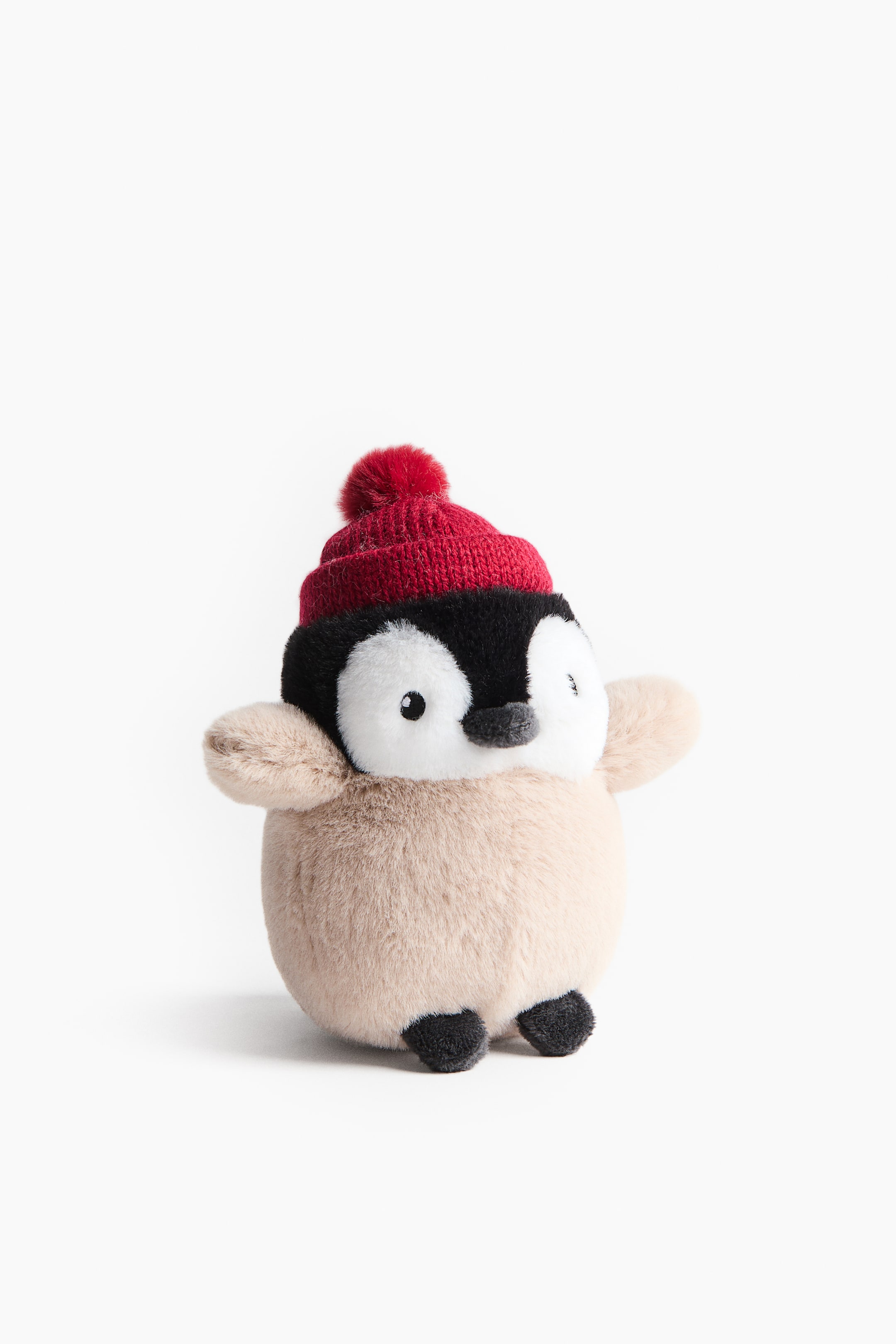 Voir une image plus grande: Une peluche de pingouin est debout, légèrement orientée vers l’avant, avec une tête noire au visage blanc, un petit bec noir et des yeux noirs. Un bonnet tricoté rouge avec un pompon orne sa tête, et son corps duveteux brun pâle laisse voir de petites nageoires brun pâle ainsi que de petits pieds noirs.