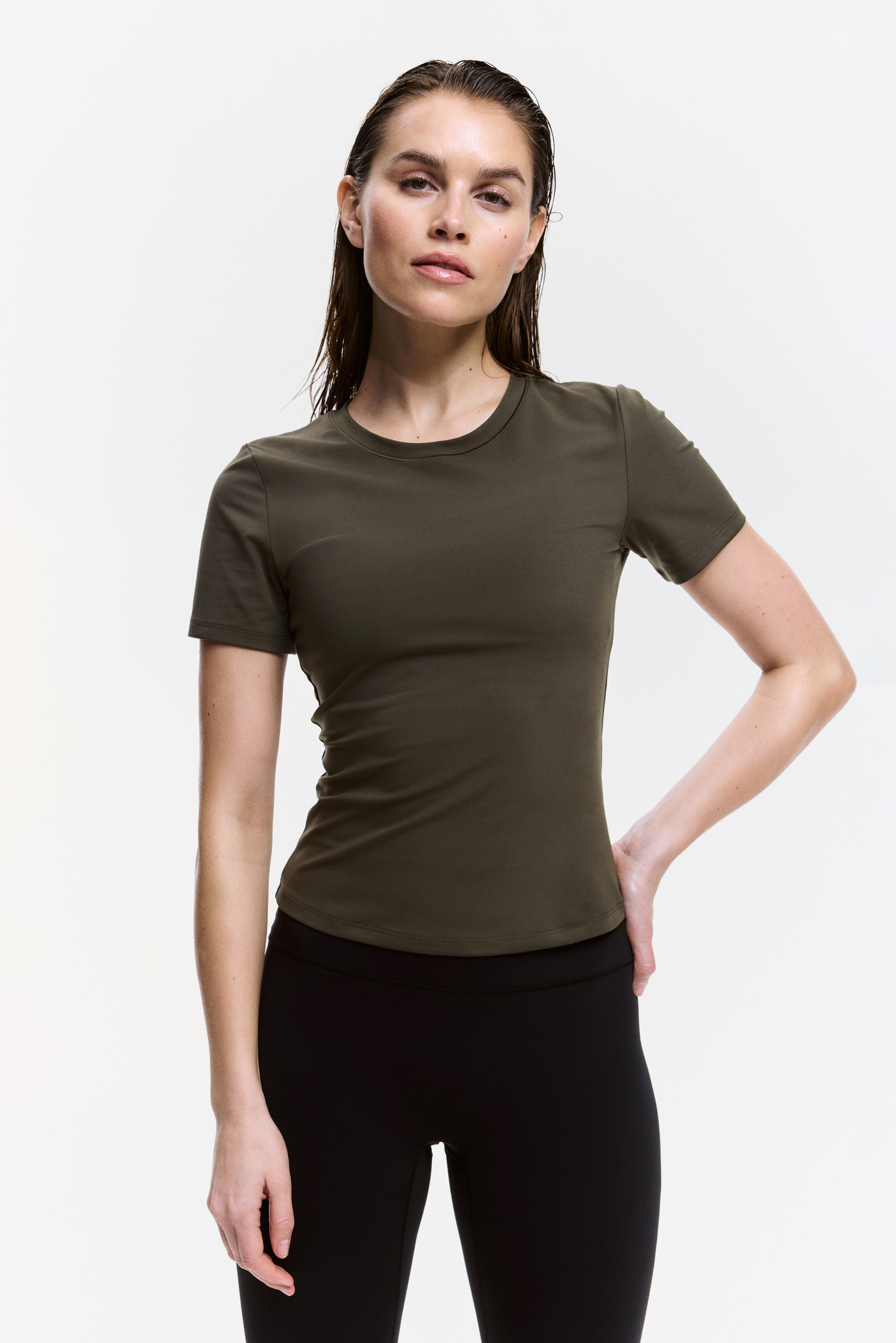 View larger image: A dark olive short-sleeved t-shirt is showcased on a person, facing forward. It features a crew neck, short sleeves, and a fitted silhouette, with its straight hemline finishing at the top of black leggings.