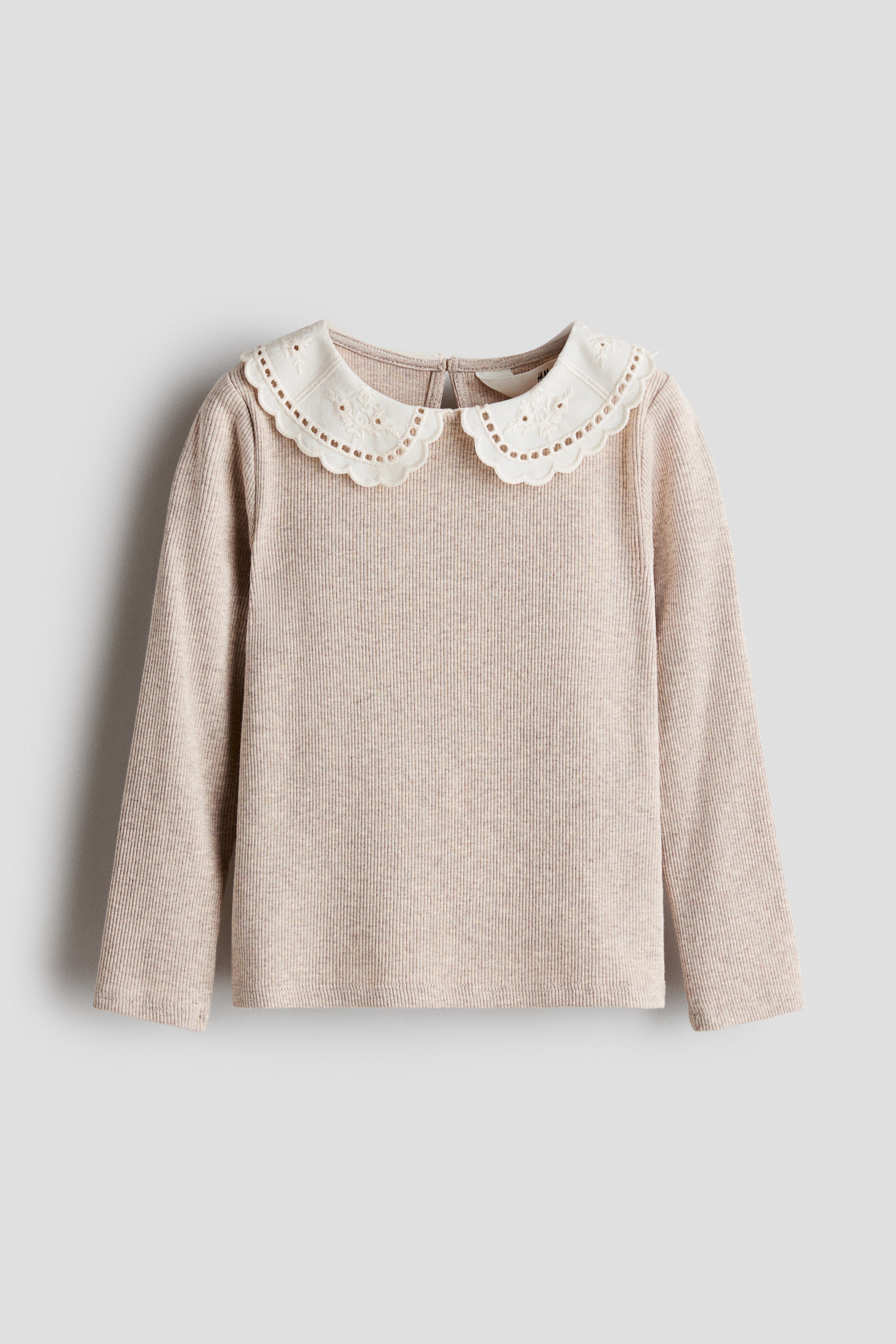 View larger image: A long-sleeve top in light beige fine ribbed jersey is shown from the front, featuring a white woven Peter Pan collar with embroidered details and scalloped edges. The top has a round neckline and a partially visible keyhole opening at the back of the neck.