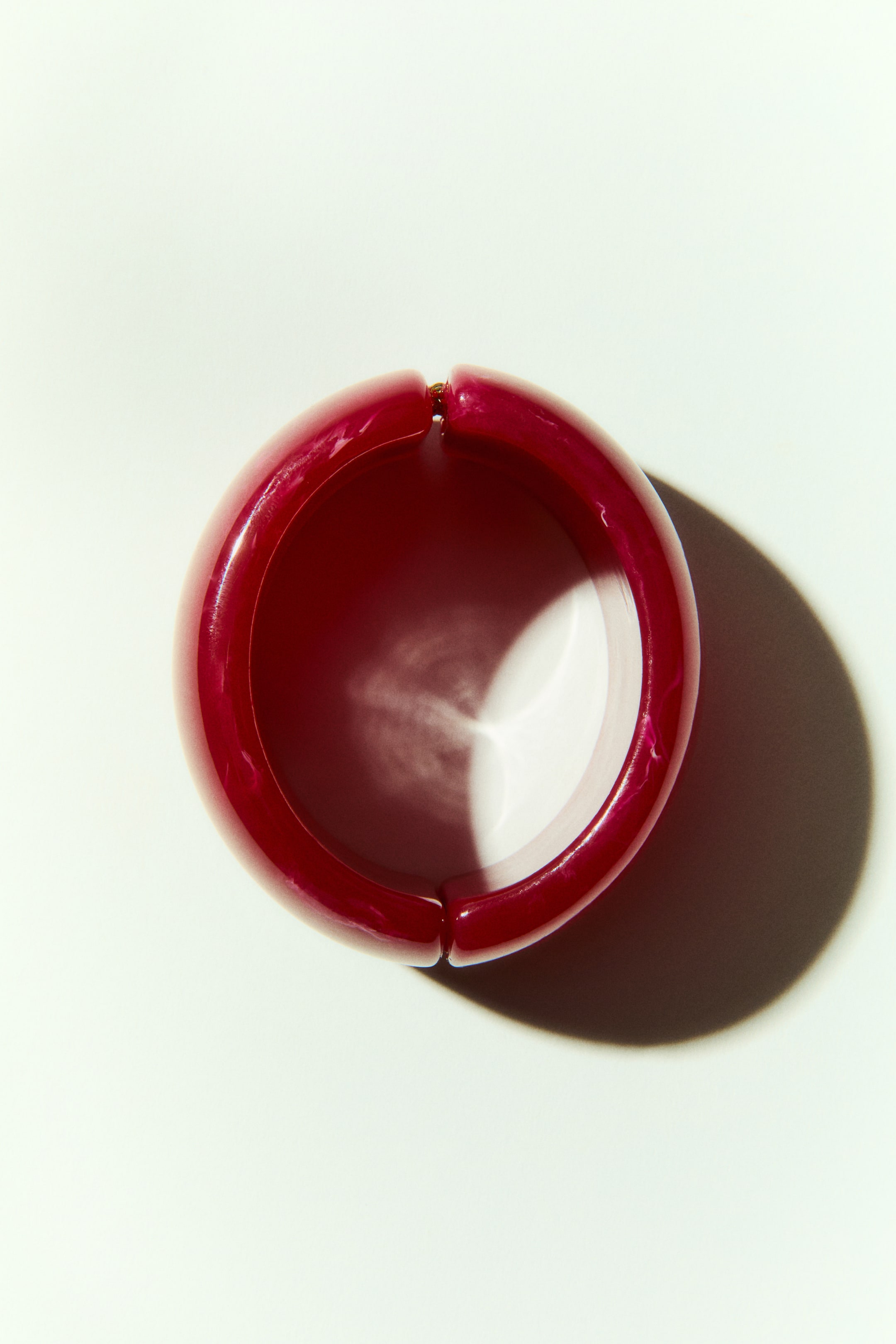 View larger image: The chunky, hinged bangle showcases a glossy, marbled deep red plastic. Its open circular design reveals a white interior, with the hinge prominently positioned at the top.