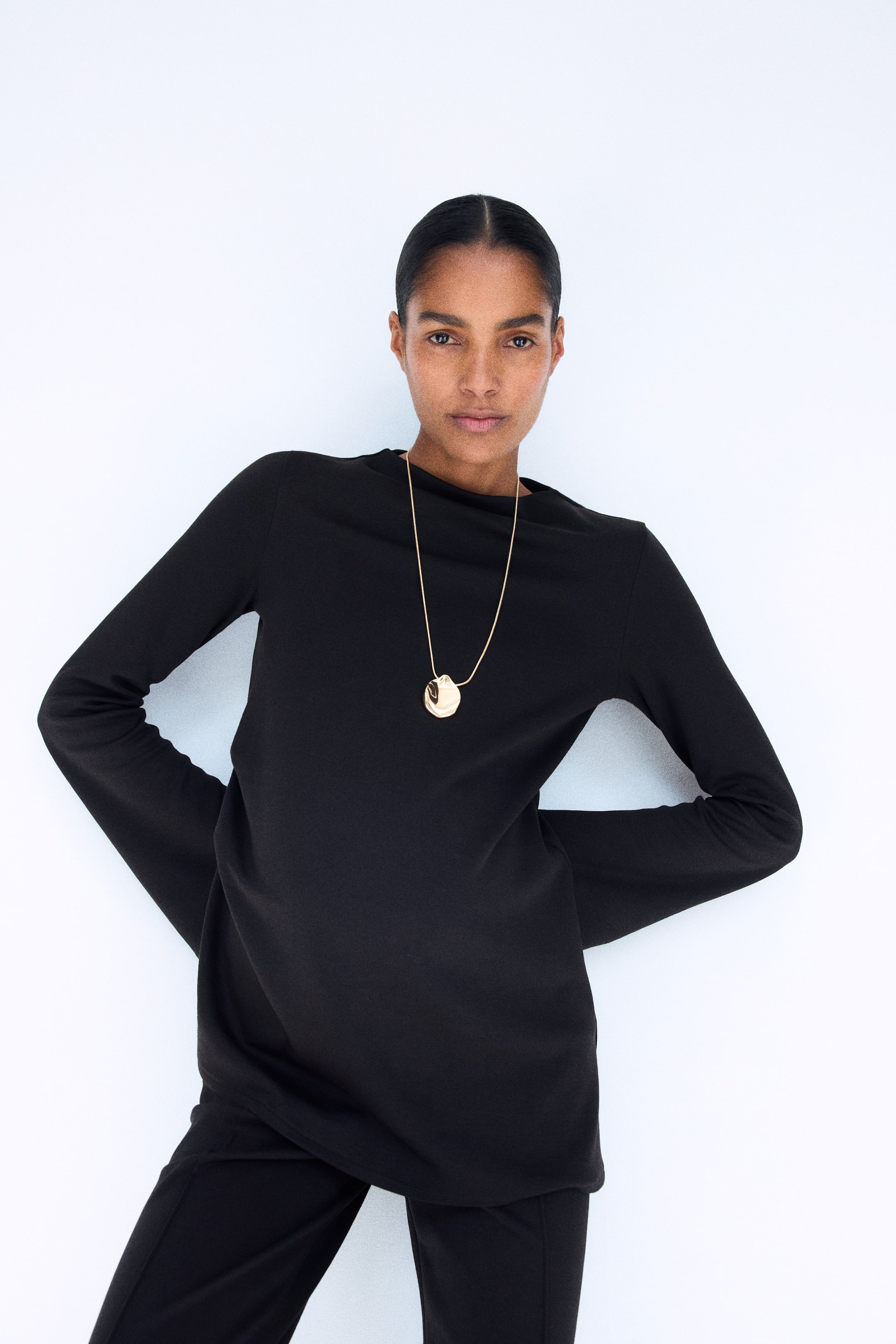 Visualizza l'immagine ingrandita: La modella indossa un completo composto da un top nero in jersey interlock e pantaloni abbinati. Il top presenta un collo alto e maniche lunghe a tromba, ed è abbinato a una collana con pendente color oro.