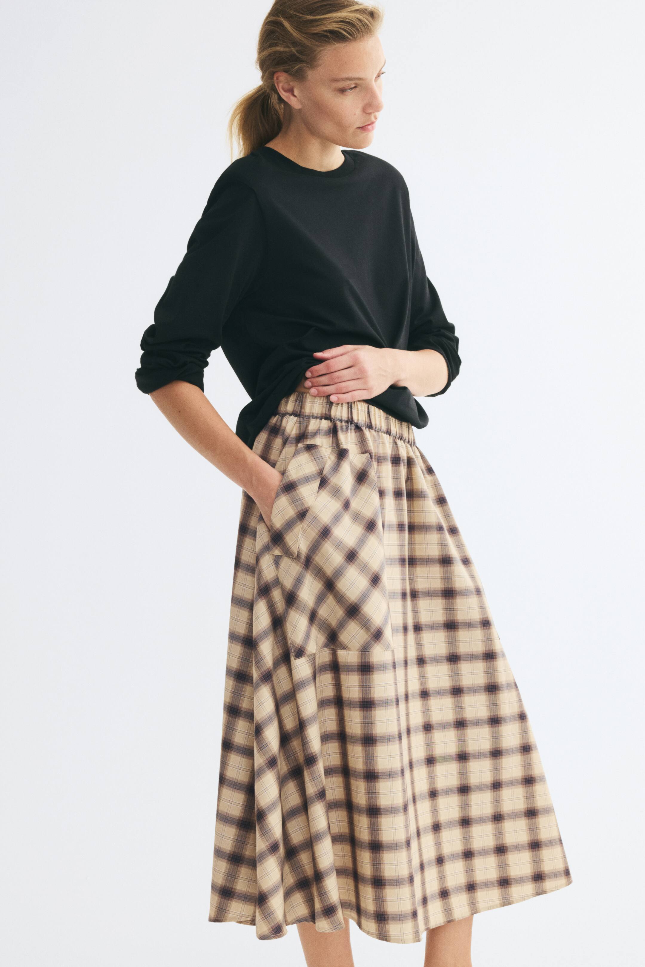 Grotere afbeelding bekijken: Een beige en bruin geruite, cirkelvormige rok tot op kuitlengte drapeert op een model dat rechtop staat en naar rechts kijkt. De rok toont een gerimpelde, elastische tailleband, die gedeeltelijk zichtbaar is onder een zwarte top met lange mouwen, en een grote opgestikte zijzak met klep.