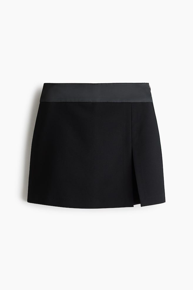 A black mini skirt is shown from the front, featuring a contrasting smooth satin waistband and a structured wrap-front design. The skirt features a vertical overlap slit on the right-hand side, creating a clean, modern silhouette. - 1
