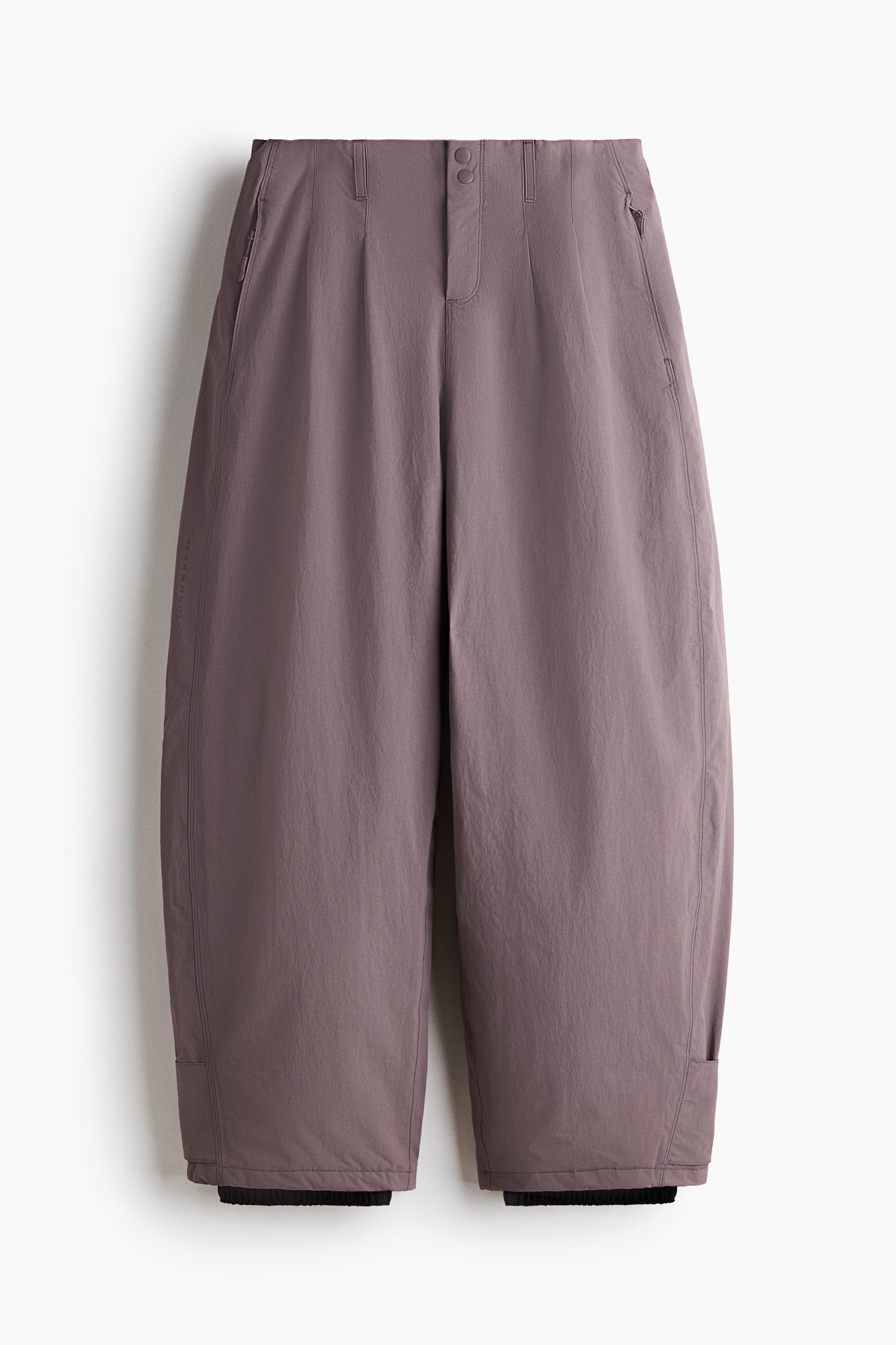 The model is wearing muted purple wide-leg ski pants with a high-waisted fit and a relaxed silhouette. The pants are styled with a black long-sleeve turtleneck, white-framed ski goggles, and black ski boots.
