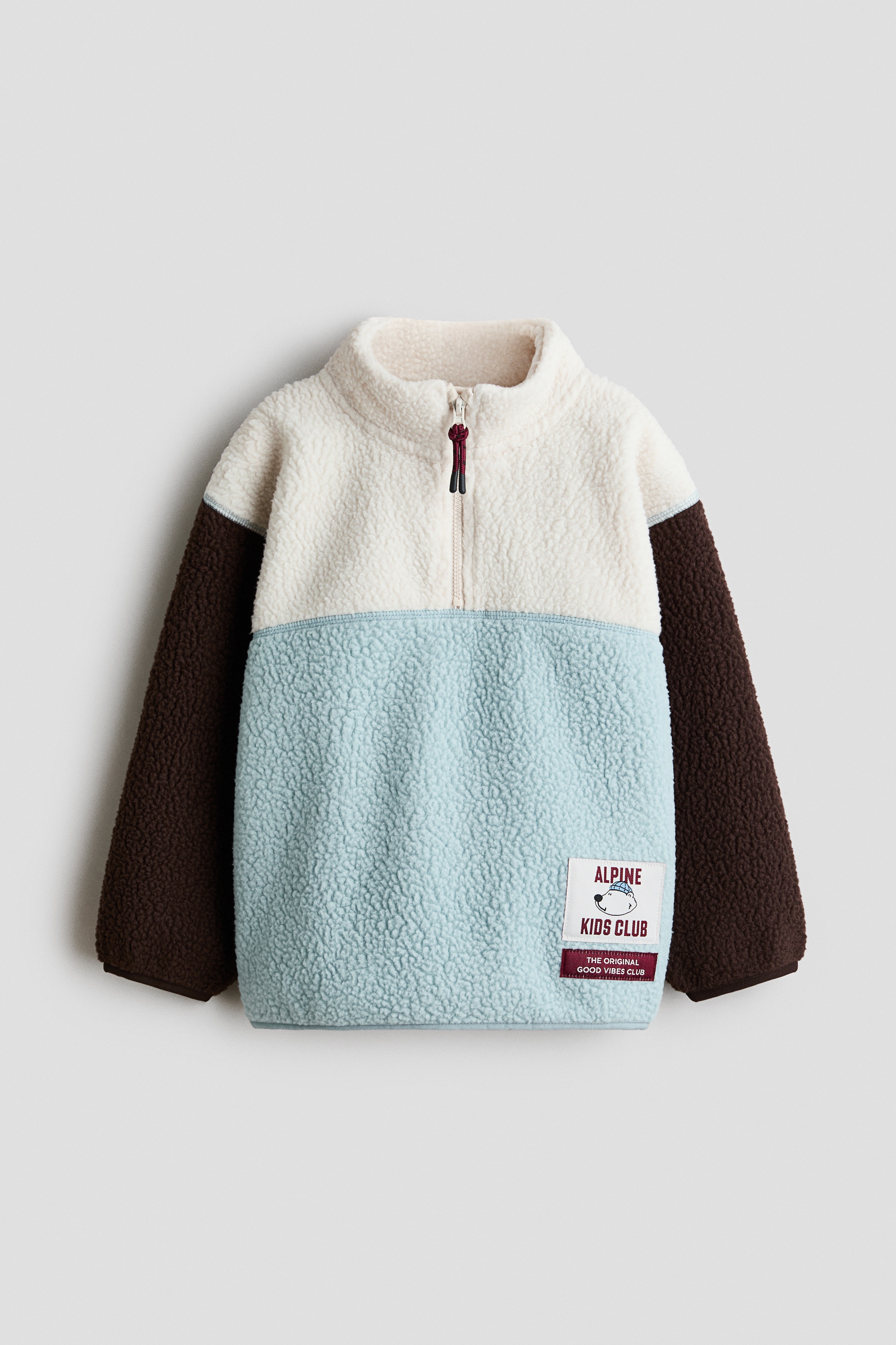 A colour-blocked teddy fleece jumper is shown from the front, featuring a cream zip-up stand collar, a light blue body, and brown sleeves with dropped shoulders. The jumper is detailed with dark jersey binding at the cuffs and hem, and a small graphic patch near the lower left.