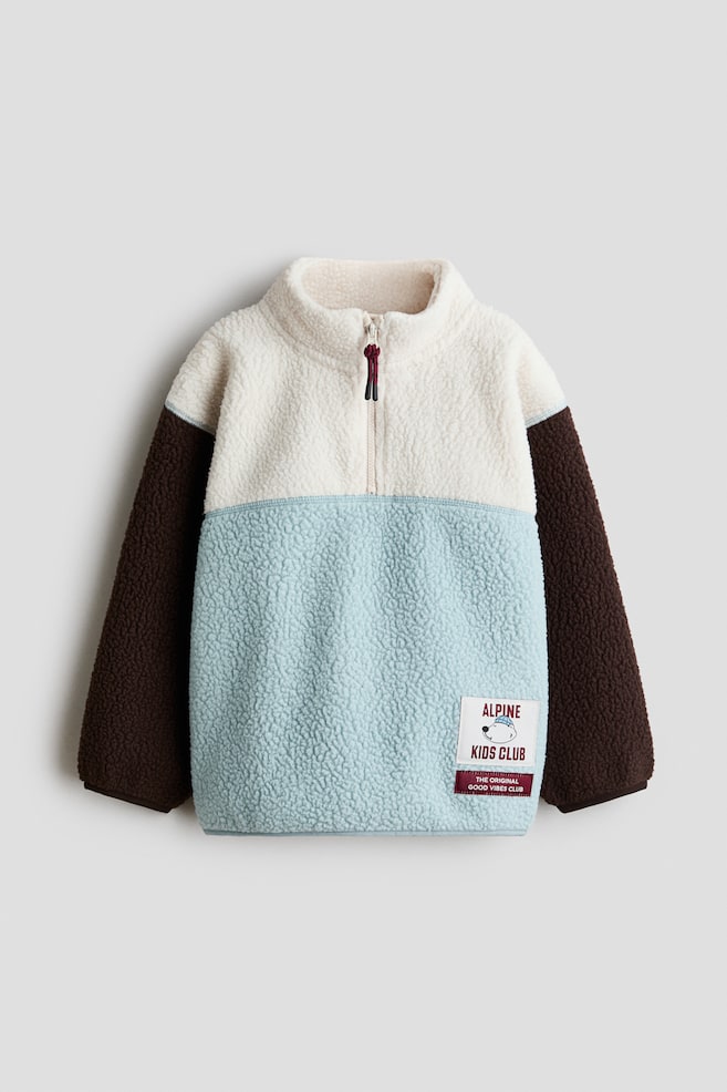 A colour-blocked teddy sweater is shown from the front, featuring an off-white upper section with a zipped stand collar, a light blue lower body, and dark brown long sleeves. The sweater has dropped shoulders, jersey binding at the cuffs and hem, and a small graphic patch at the lower right. - 1