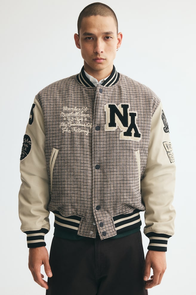 A baseball jacket featuring a brown-and-beige houndstooth woven body and cream sleeves with decorative patches is worn by the model. The jacket is designed with a large "NY" appliqué on the chest, a press-stud front fastening, and striped ribbed trim on the collar, cuffs, and hem. It is styled over a white collared shirt and paired with dark trousers. - 1