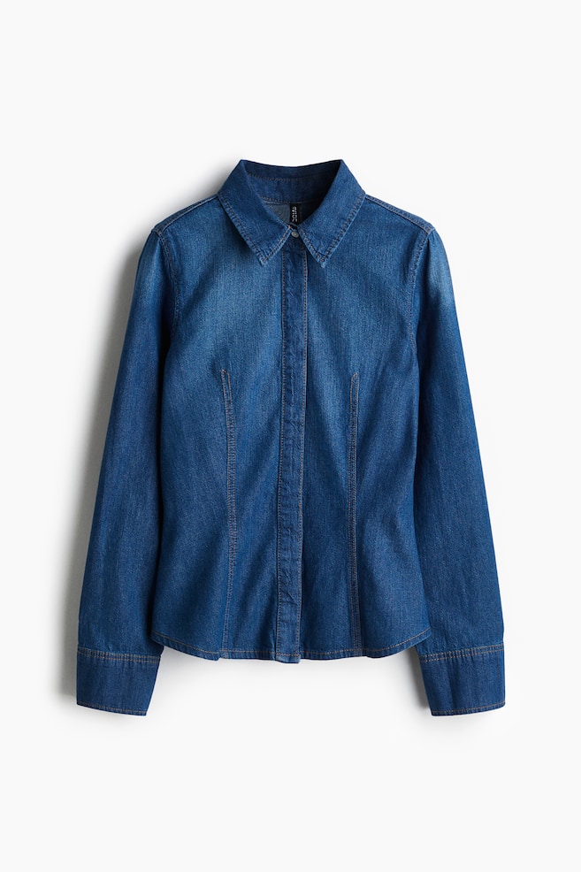 Eine Vorderansicht eines blauen Jeanshemds wird präsentiert, mit einem spitzen Kragen, einer Knopfleiste vorne und langen Ärmeln mit geknöpften Manschetten. Das Hemd zeigt einen gewaschenen Denim-Effekt mit helleren Bereichen an den Schultern und der Brust sowie sichtbaren Kontrastnähten, die vertikale Nähte für ein tailliertes Aussehen umreißen. - 1